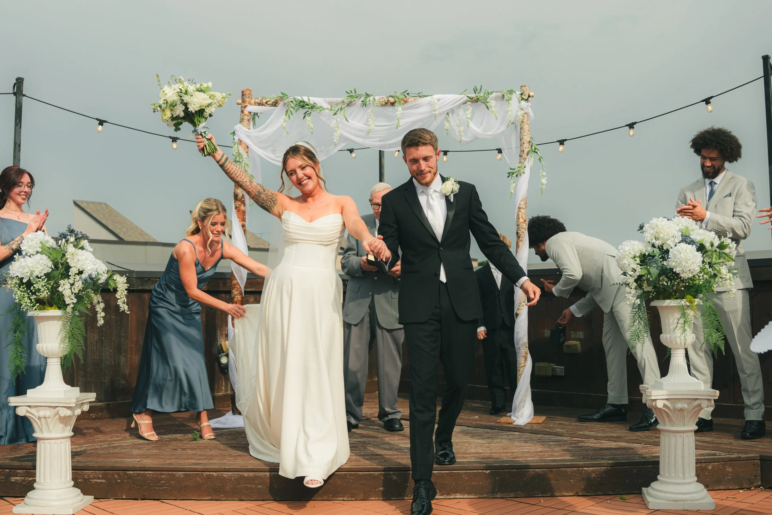 A wedding ceremony on a rooftop with a female bride in a white dress and a male groom in a black suit holding hands under a decorated arch. They are surrounded by friends and family, some clapping, with flower arrangements and string lights.