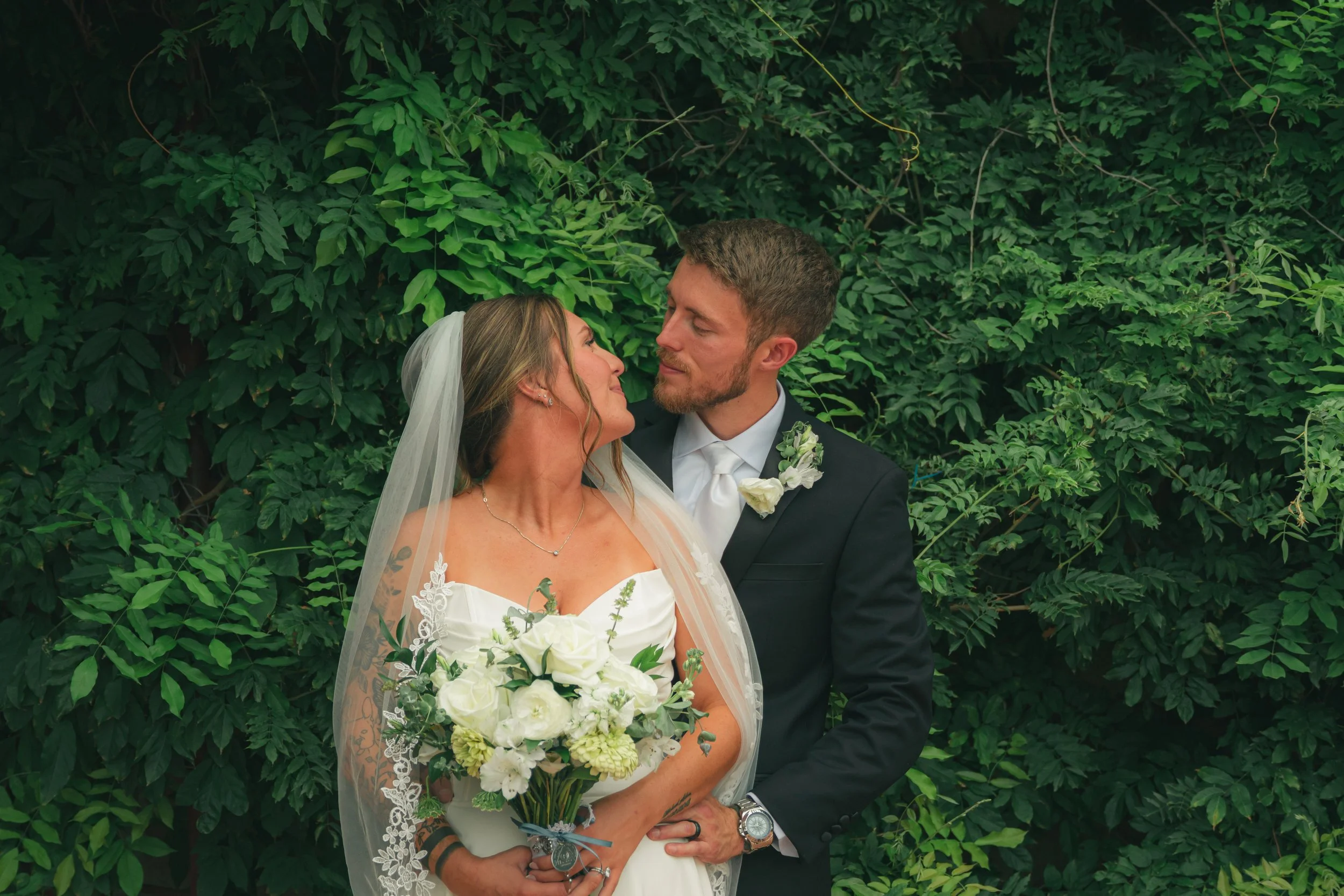A bride and groom embrace in front of green foliage; the bride holds a bouquet of white flowers and is wearing a lace wedding dress with a veil, while the groom is wearing a black suit and white shirt.