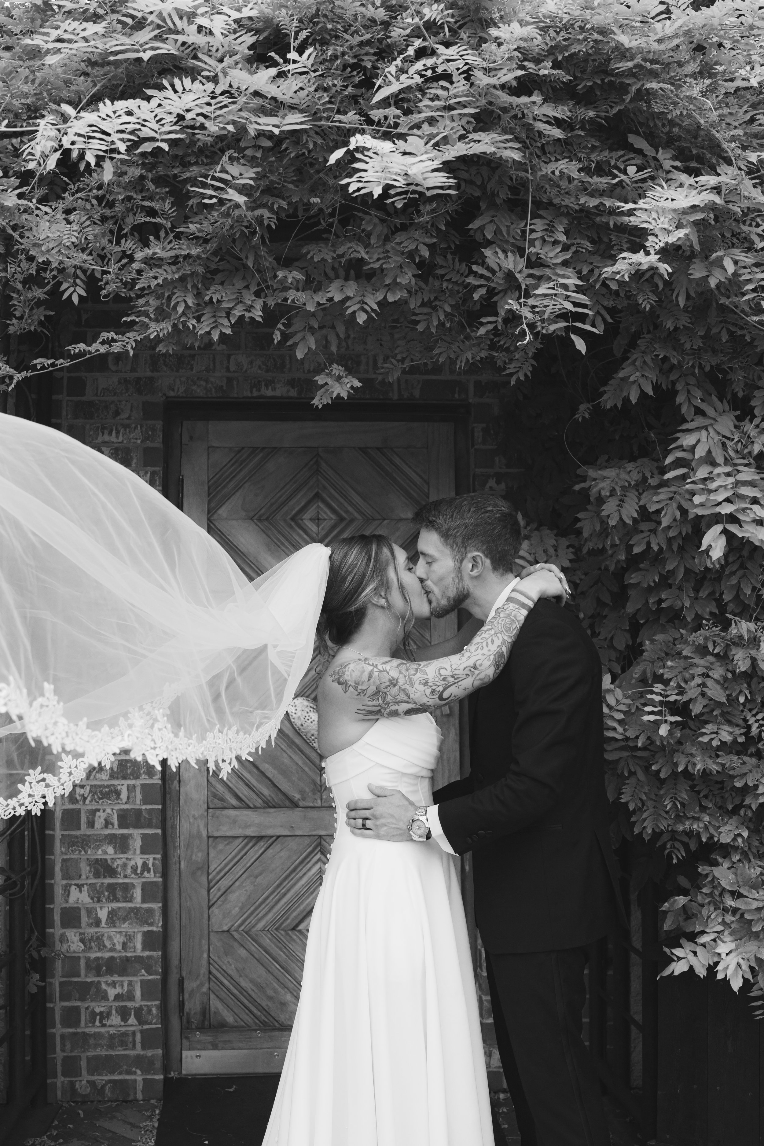 A bride and groom kissing outdoors in front of a wooden door surrounded by lush leafy plants, with part of a lace veil visible on the bride's head.