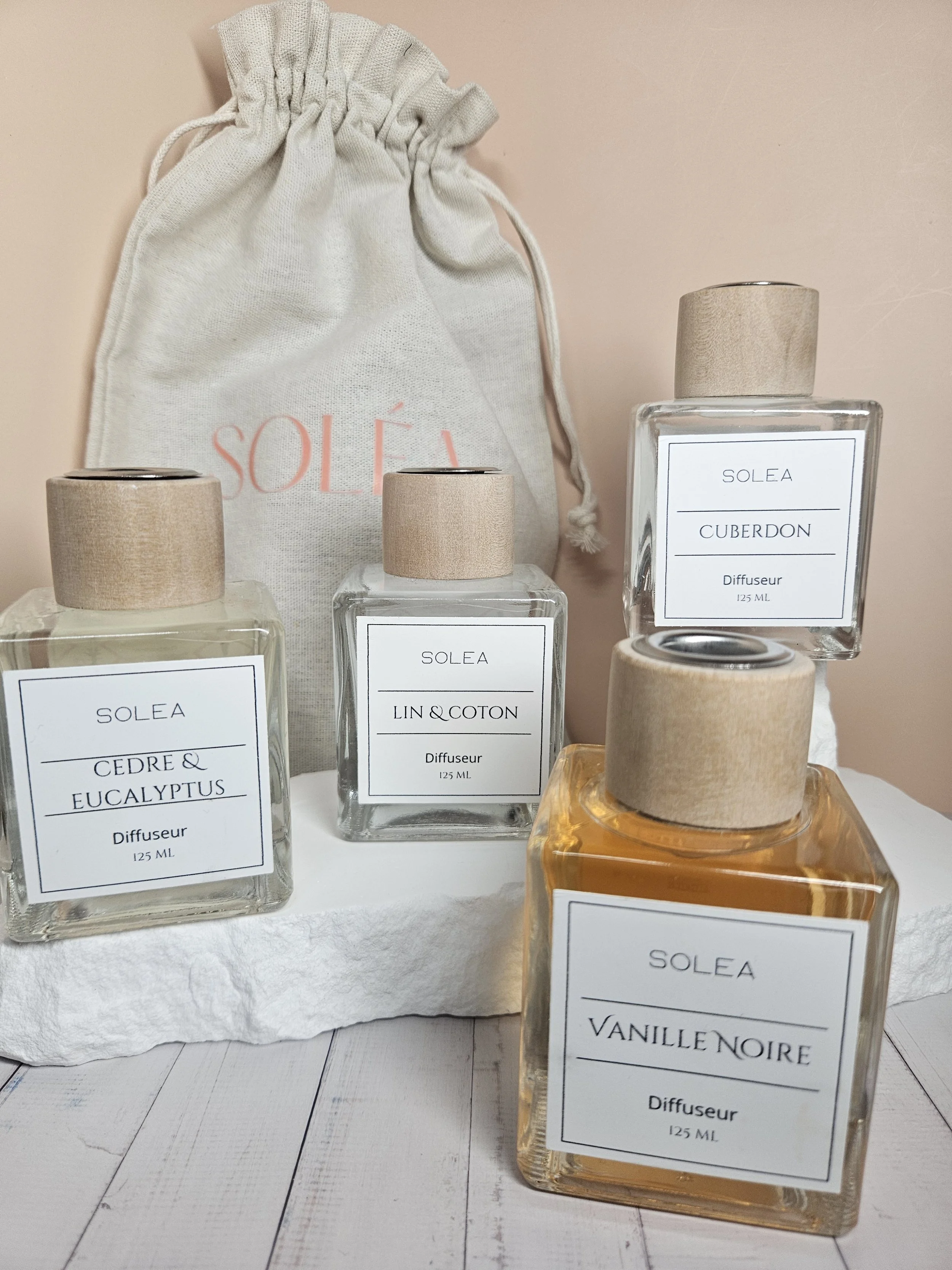Quatre bouteilles de parfum de la marque SOLEA avec différentes senteurs et un sac en tissu beige avec le logo SOLEA en rose, sur une surface blanche et un fond beige clair.
