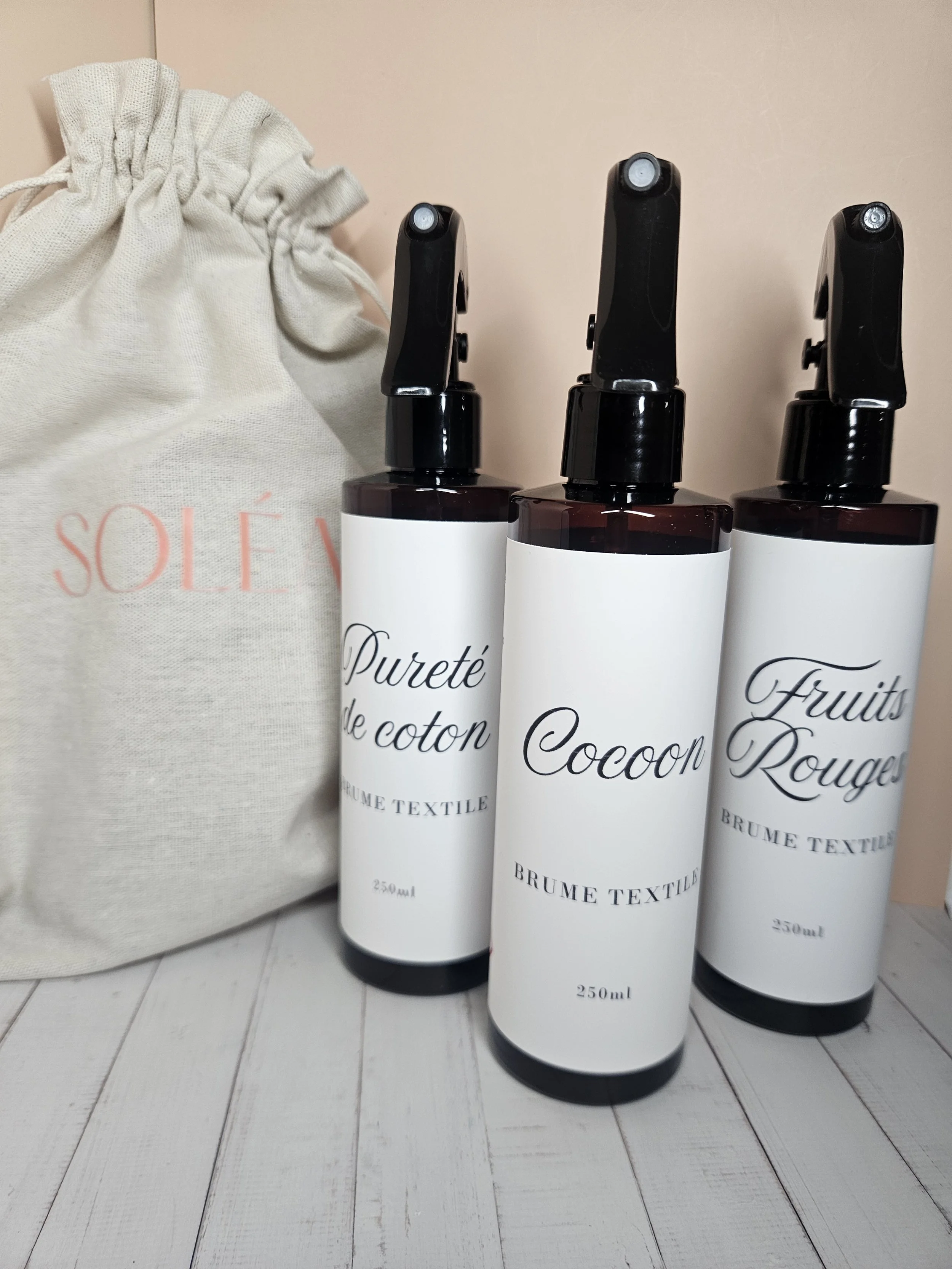 Trois bouteilles avec diffuseur spray, étiquettes indiquant 'Pureté de coton', 'Cocoon' et 'Fruits Rouges', et un sac en tissu beige avec le mot 'SOLÉV' en lettres roses à l'arrière.
