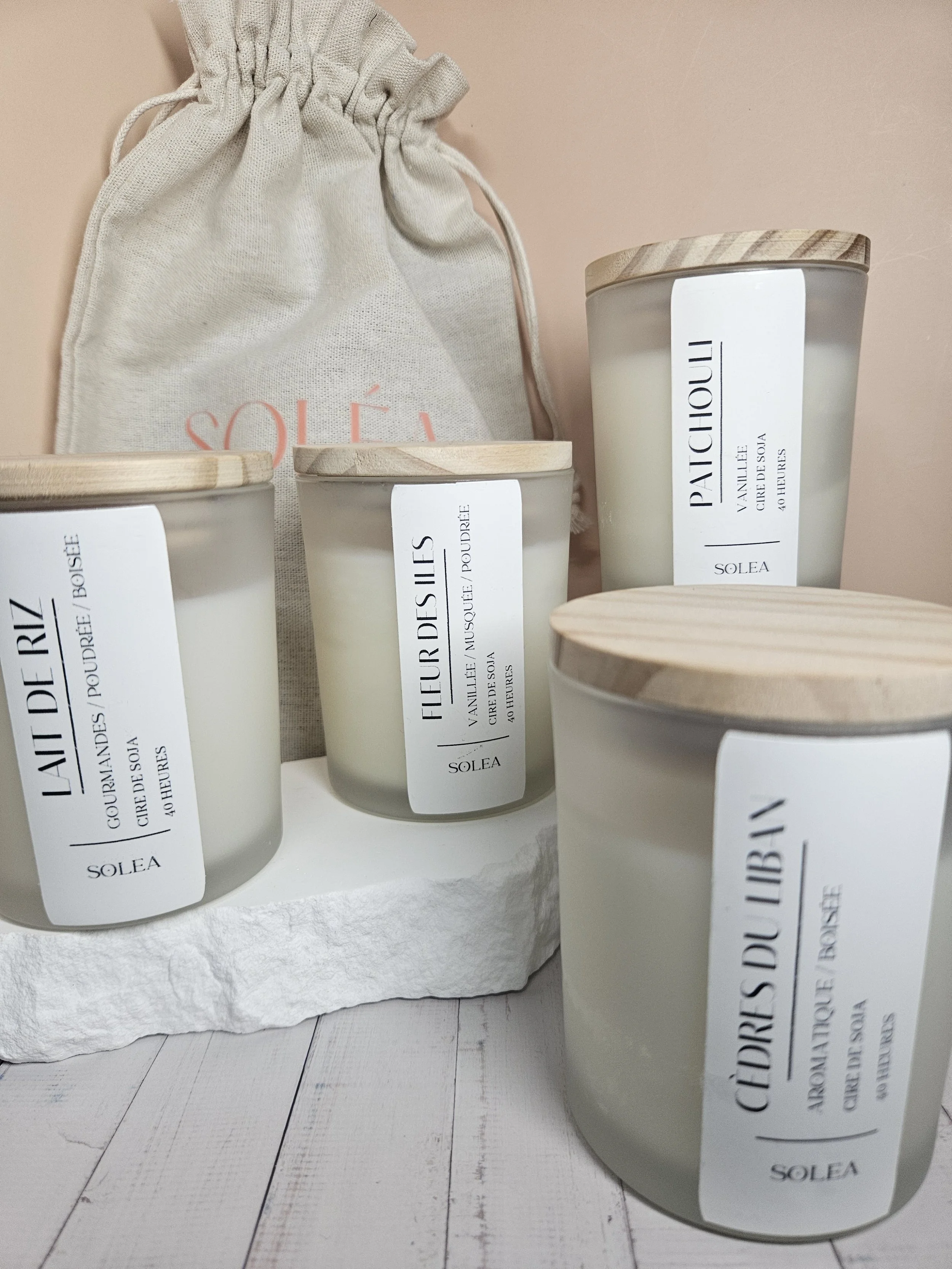 Bougies parfumées dans des pots en verre avec étiquettes blanches et couvercles en bois, placées sur une plateforme en pierre face à un sac en coton beige. Fonds beige avec sol en bois clair.