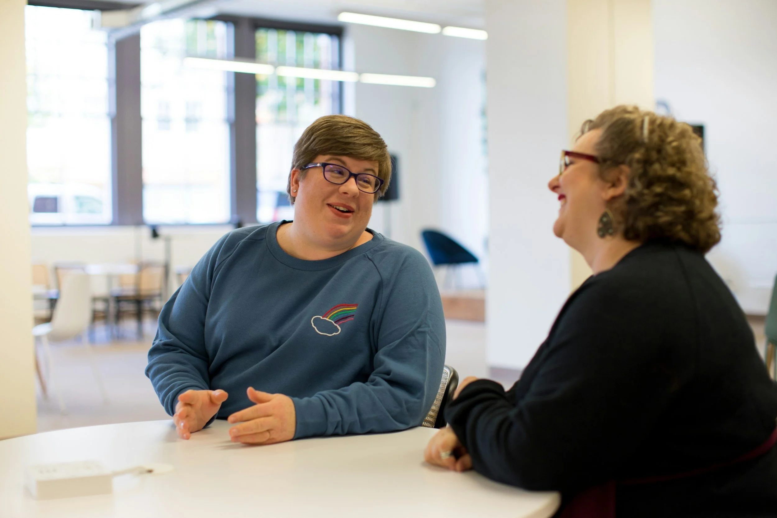 Woman in blue top with a rainbow and cloud on it and short hair and glasses talking to another woman with short curly hair and glasses in a black jumper sitting at a desk in an office near a window
