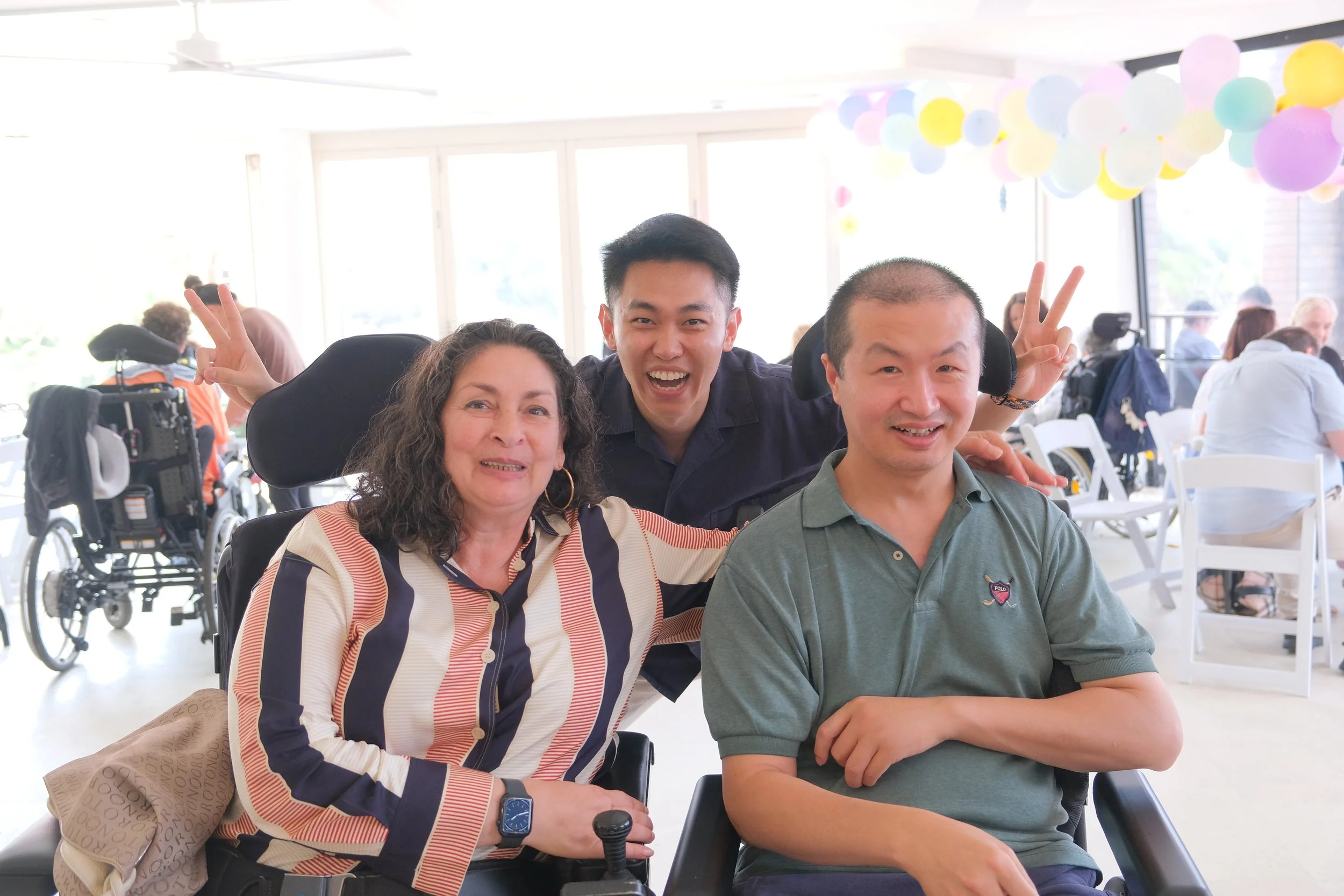 A group of people smiling and enjoying time together in a community setting, including a Mandarin-speaking support worker and a person using a wheelchair.