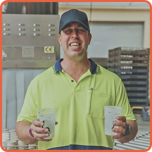 a man in a fluro workers top going back to work after being assisted by icare and cca holding two cups, wearing a hat in a factory setting