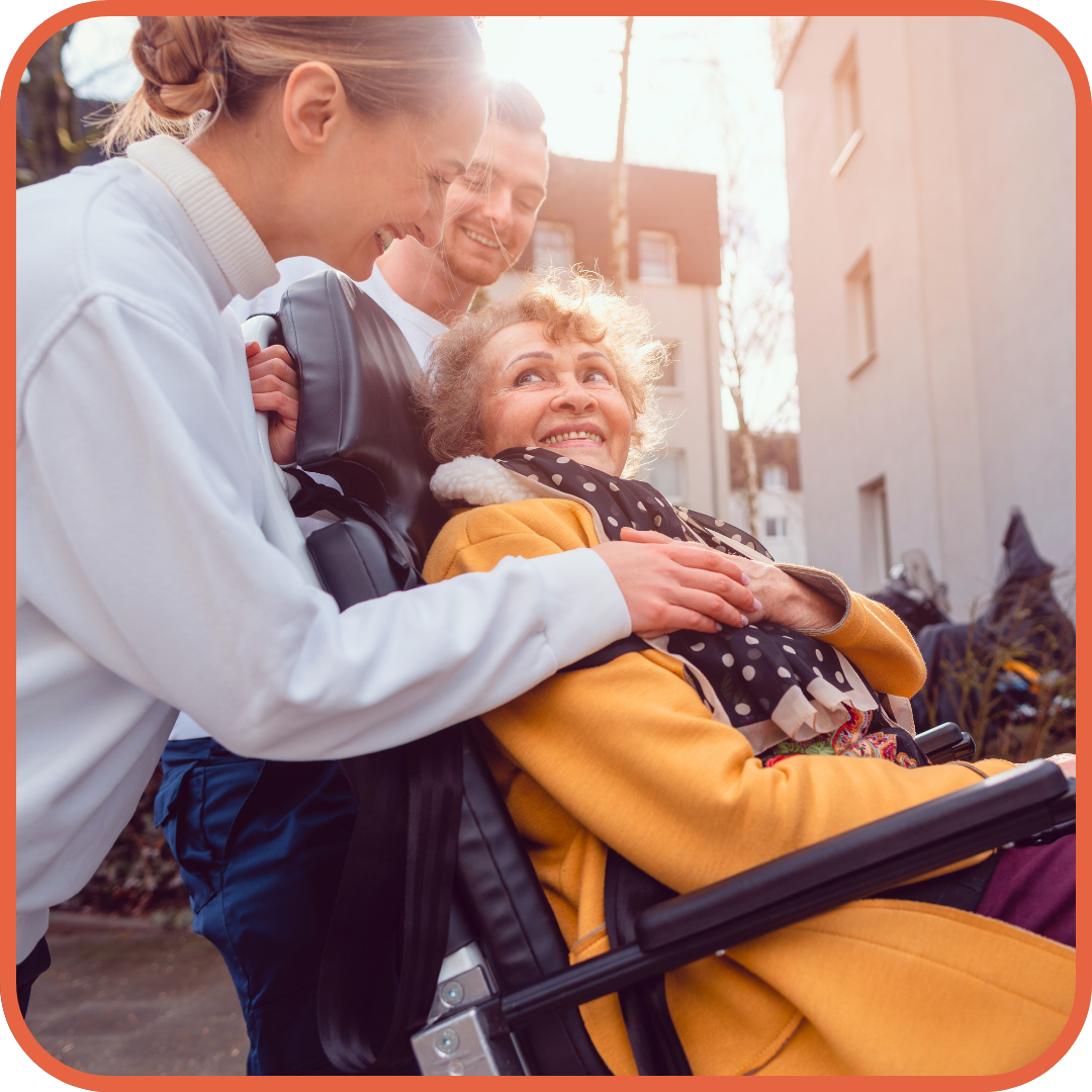 an older woman living with disability is in a wheelchair wearing a yellow jacket and dark blue scarf being supported by a smiling blonde haired woman with a bun in her hair and apartments in background
