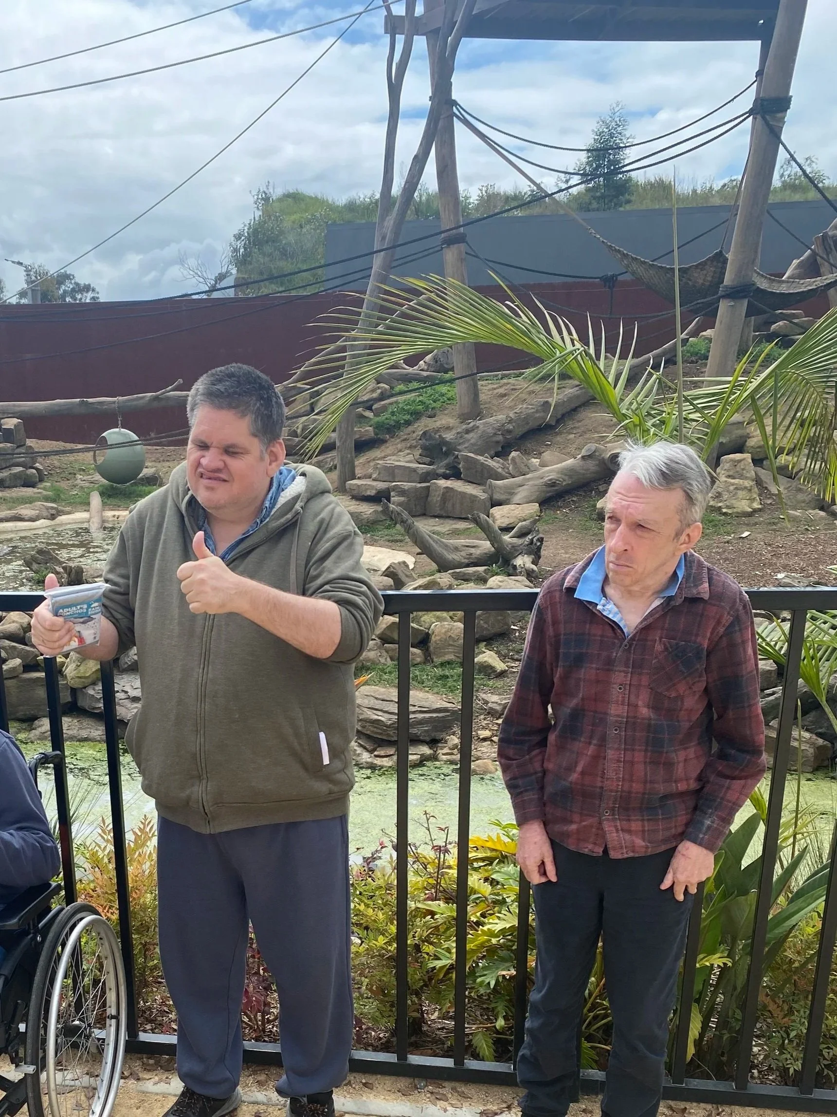 two people outdoors in front of a zoo enclosure, with one person is giving a thumbs-up sign. There are logs, rocks, and plants in the background.