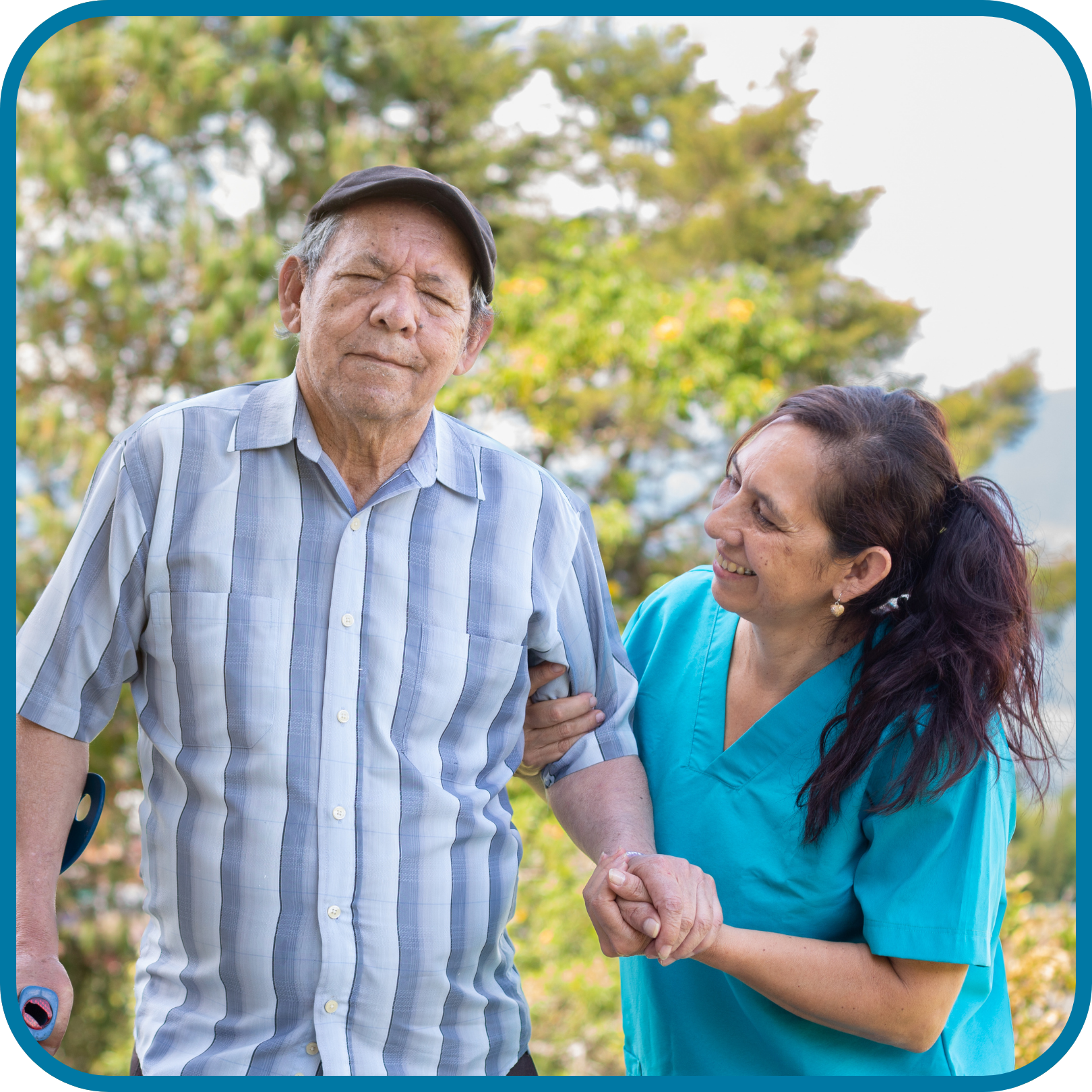 A older man living with a disability wearing a blue striped shirt and grey hat is being helped by a woman with long dark hair and a blue top walking in a park with trees in the background