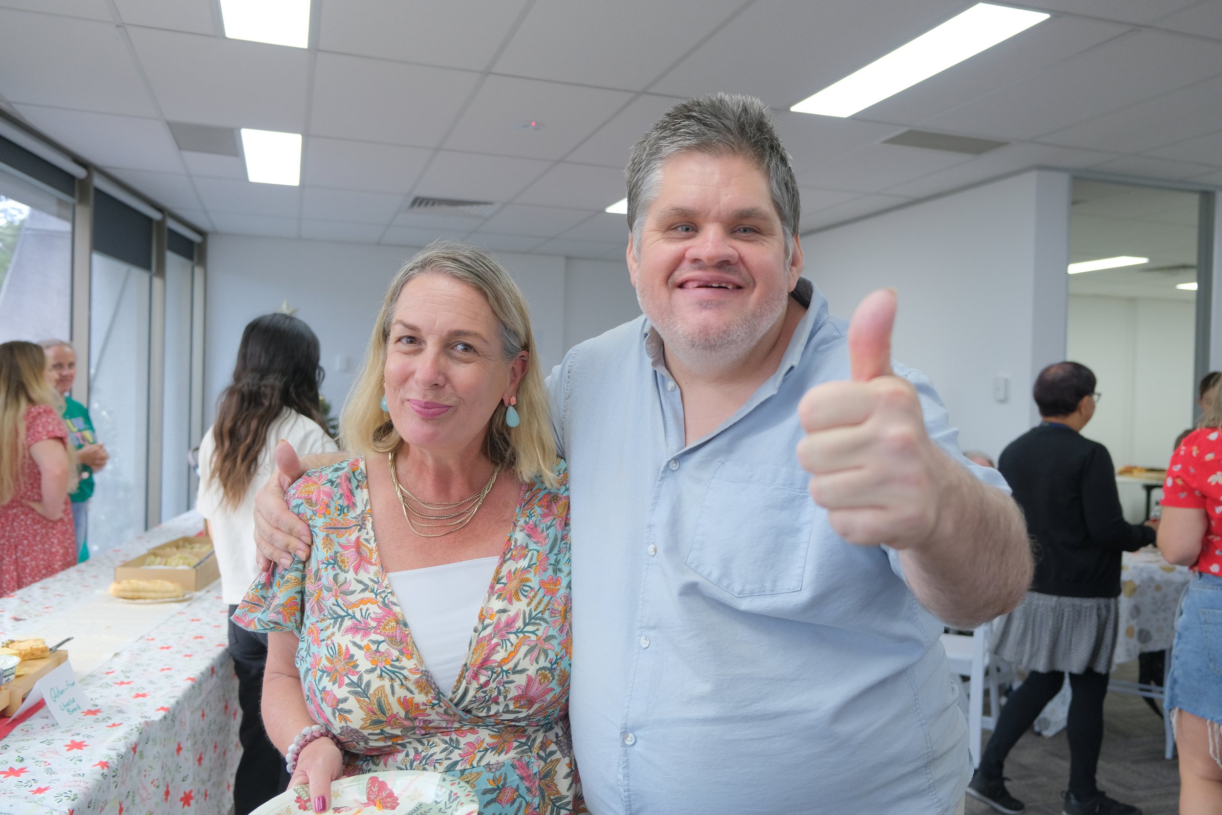A man giving thumbs up wearing a blue shirt and smiling standing with his arm around the shoulder of a lady with blonde hair in a long bob wearing a flower patterned dress holding a plate in an office in Parramatta.
