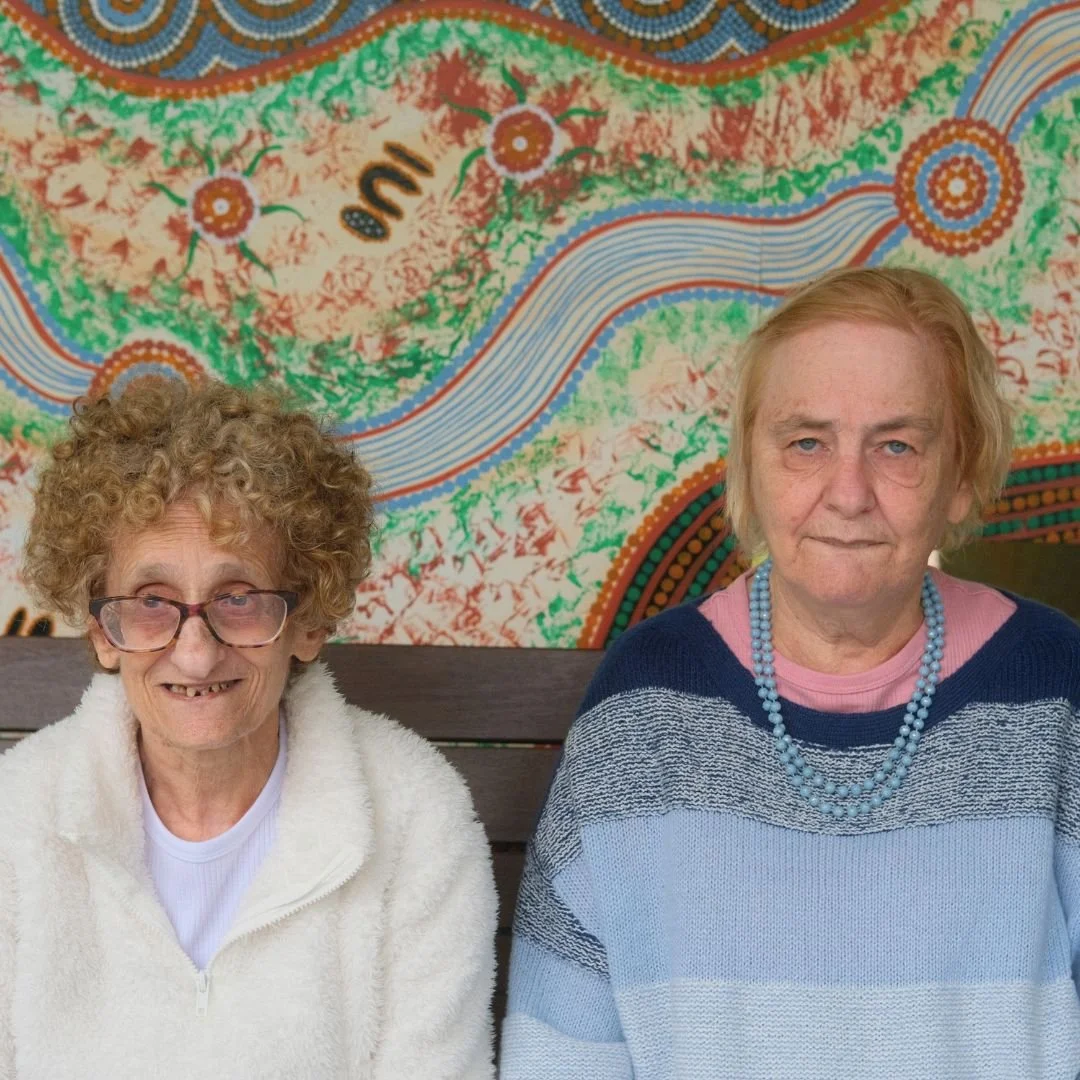 Two women smiling at the camera, seated together in front of an Aboriginal artwork.