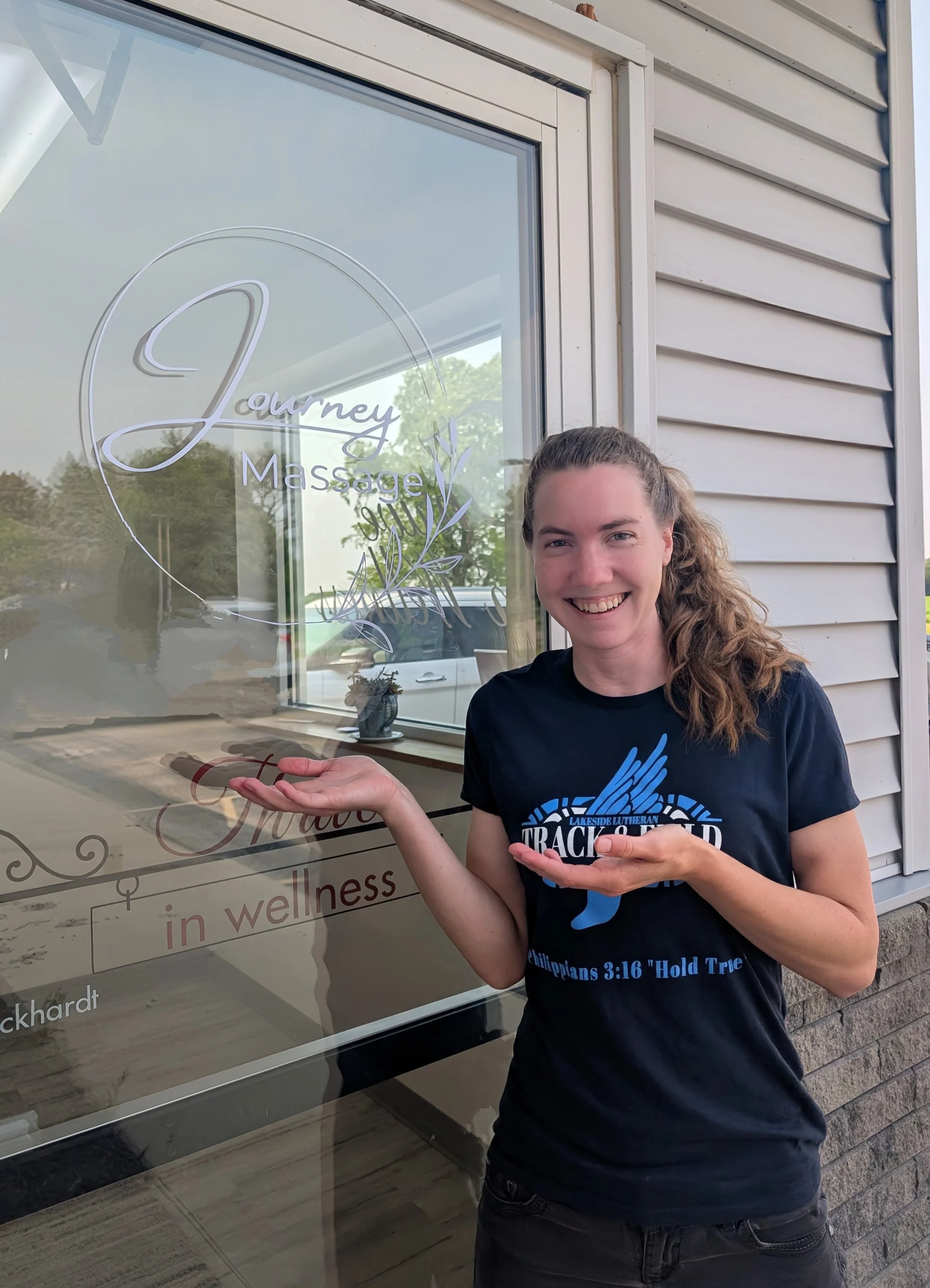 Massage therapist Danielle stands by glass door featuring a white vinyl sign for Journey Massage