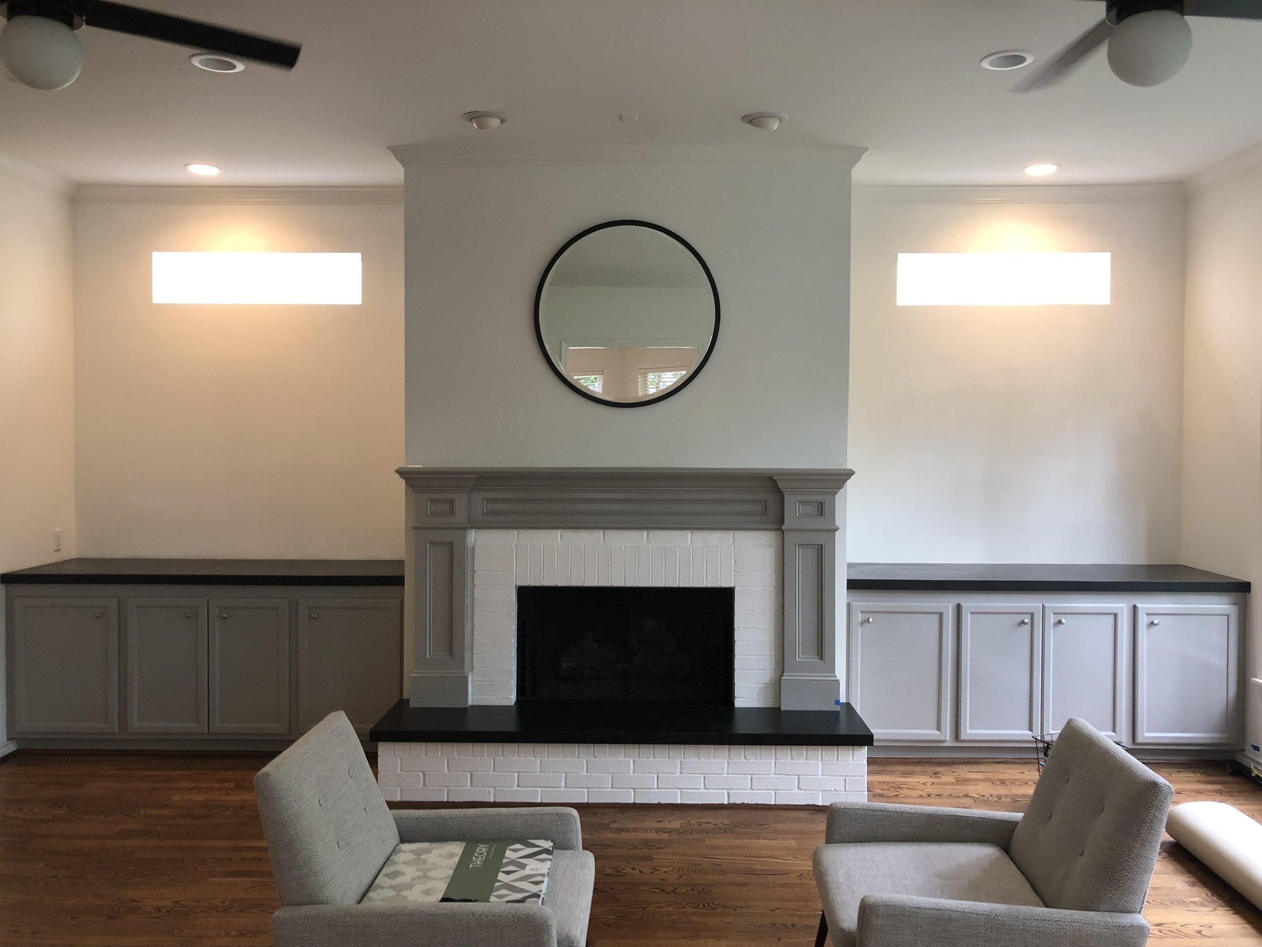 Living room interior with a white brick fireplace, gray mantel, round mirror above, gray cabinets on each side, two modern chairs, and hardwood flooring.