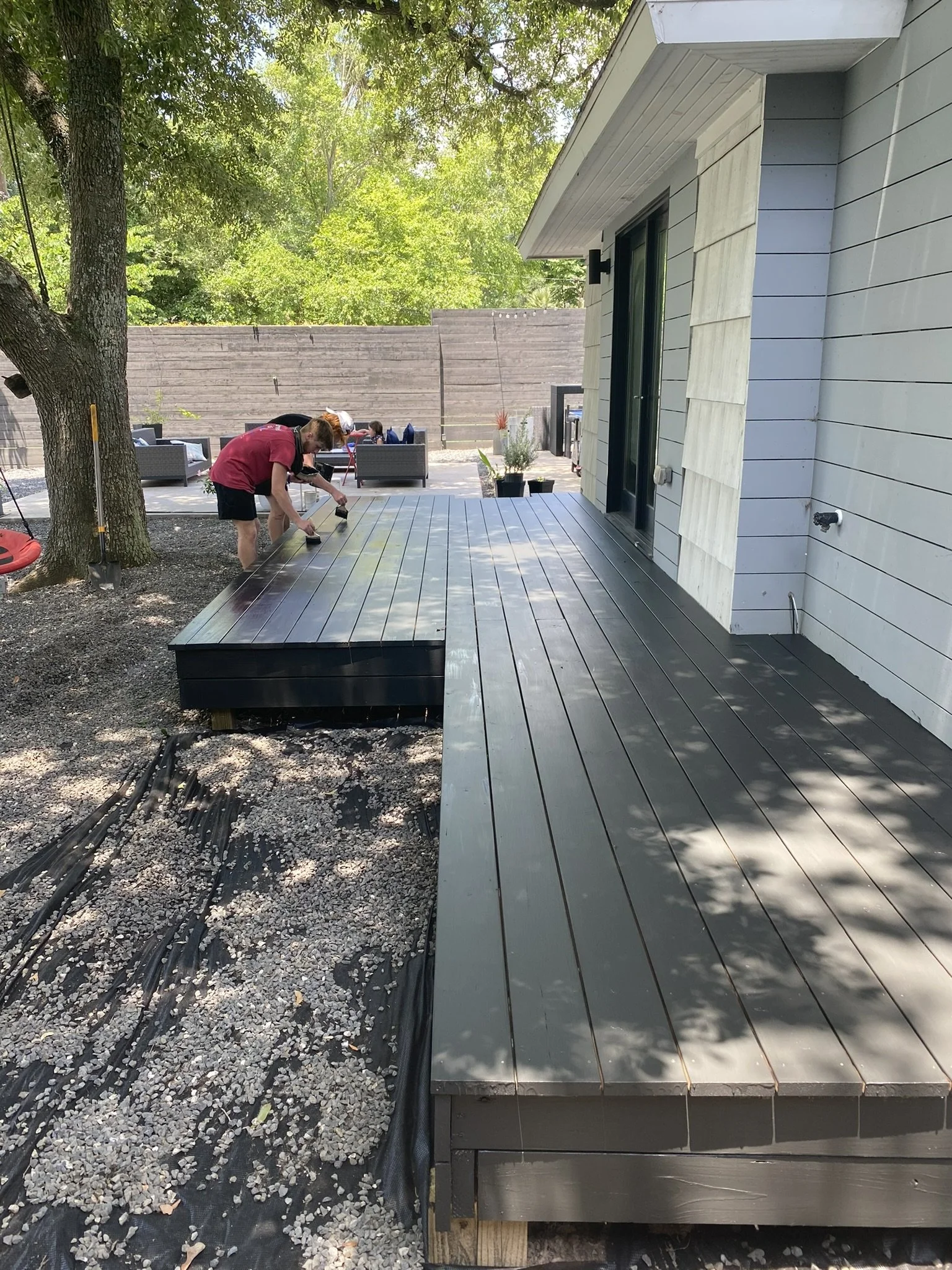 Person painting a wooden deck next to a house in a backyard with trees and outdoor furniture.