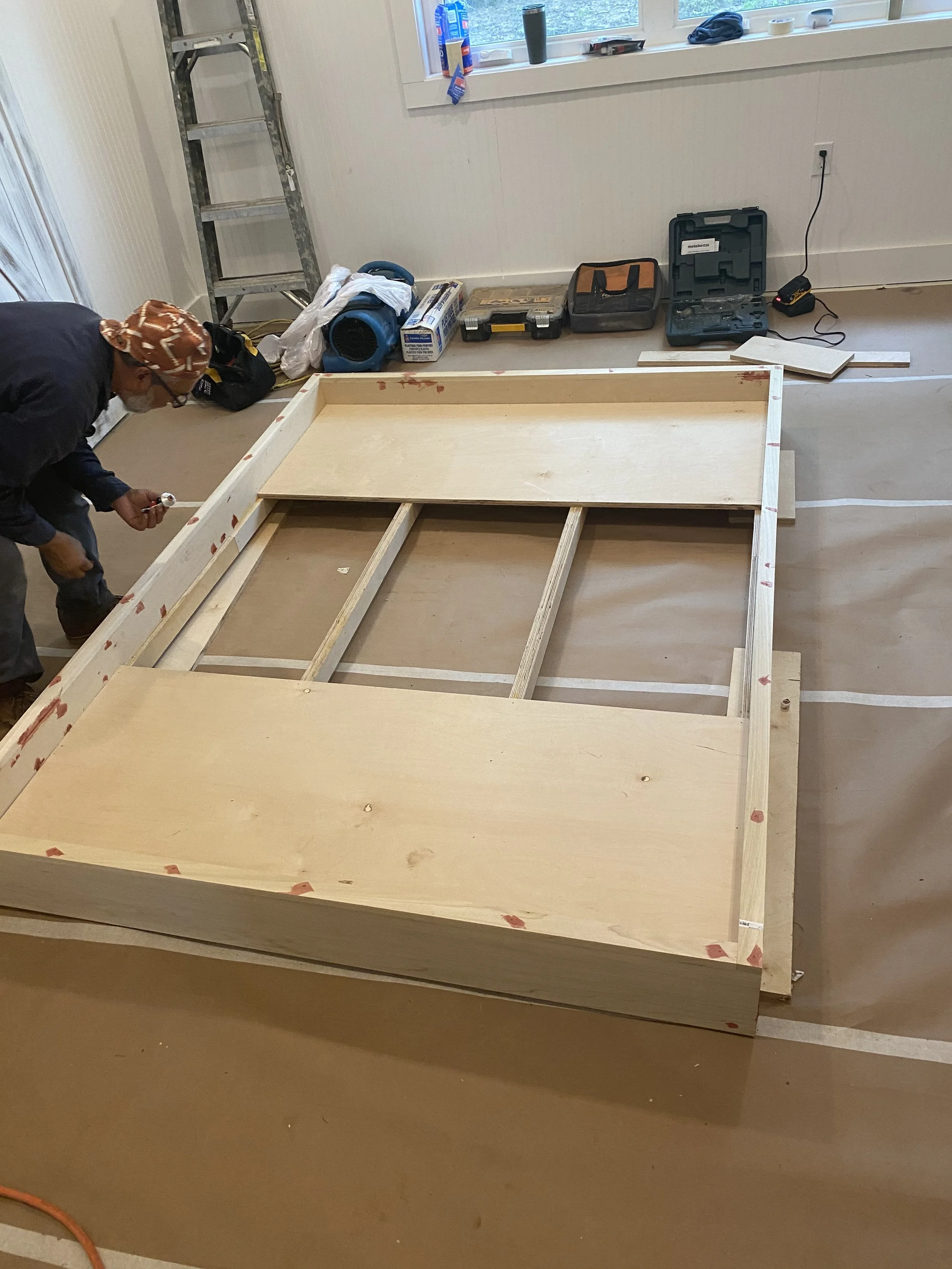 Person working on a wooden furniture piece in a room with tools and materials around, ladder in the background.