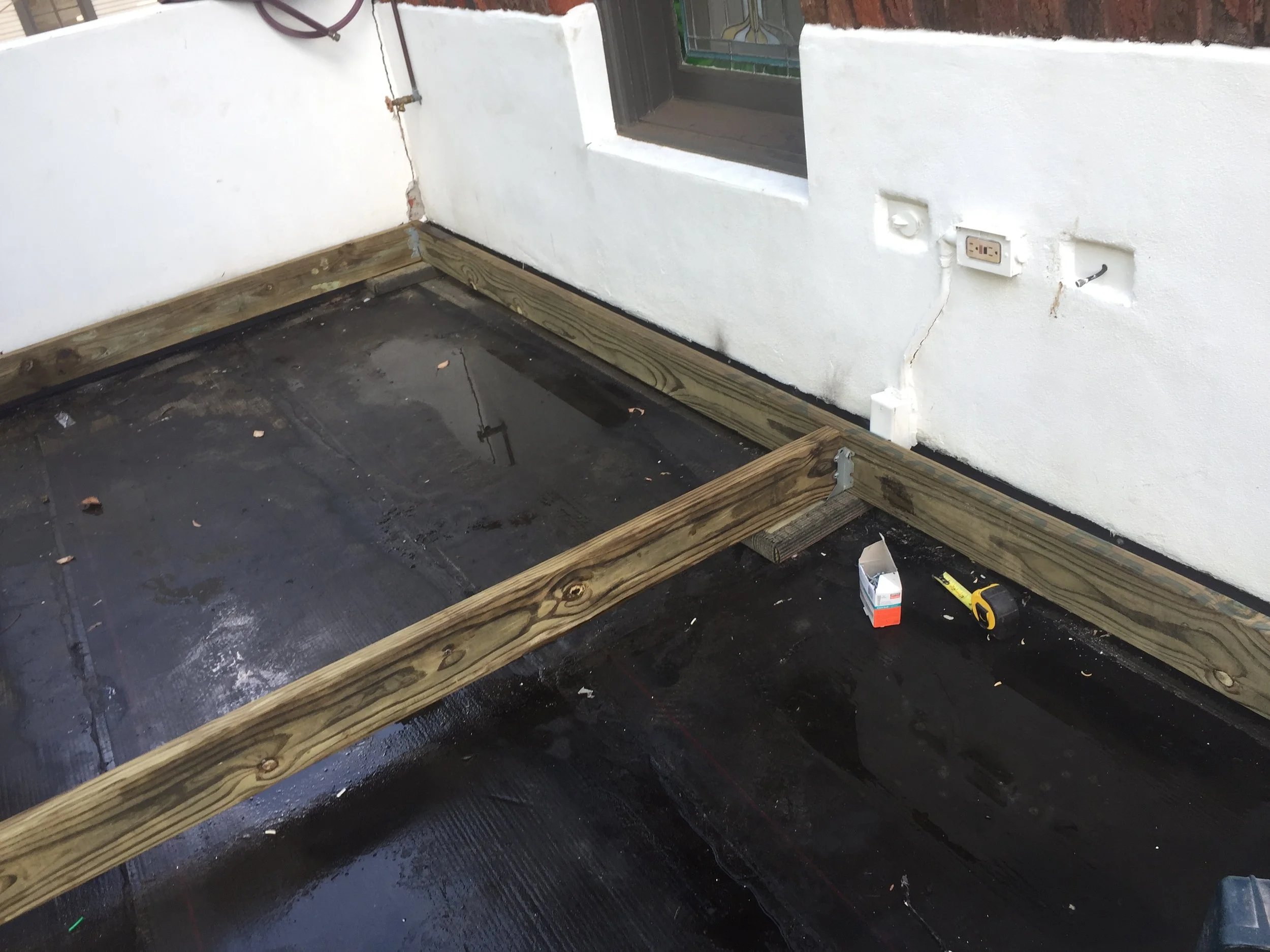 Wooden framework on a rooftop deck under construction, with a tape measure, wet surface, electrical outlet, and tools present.