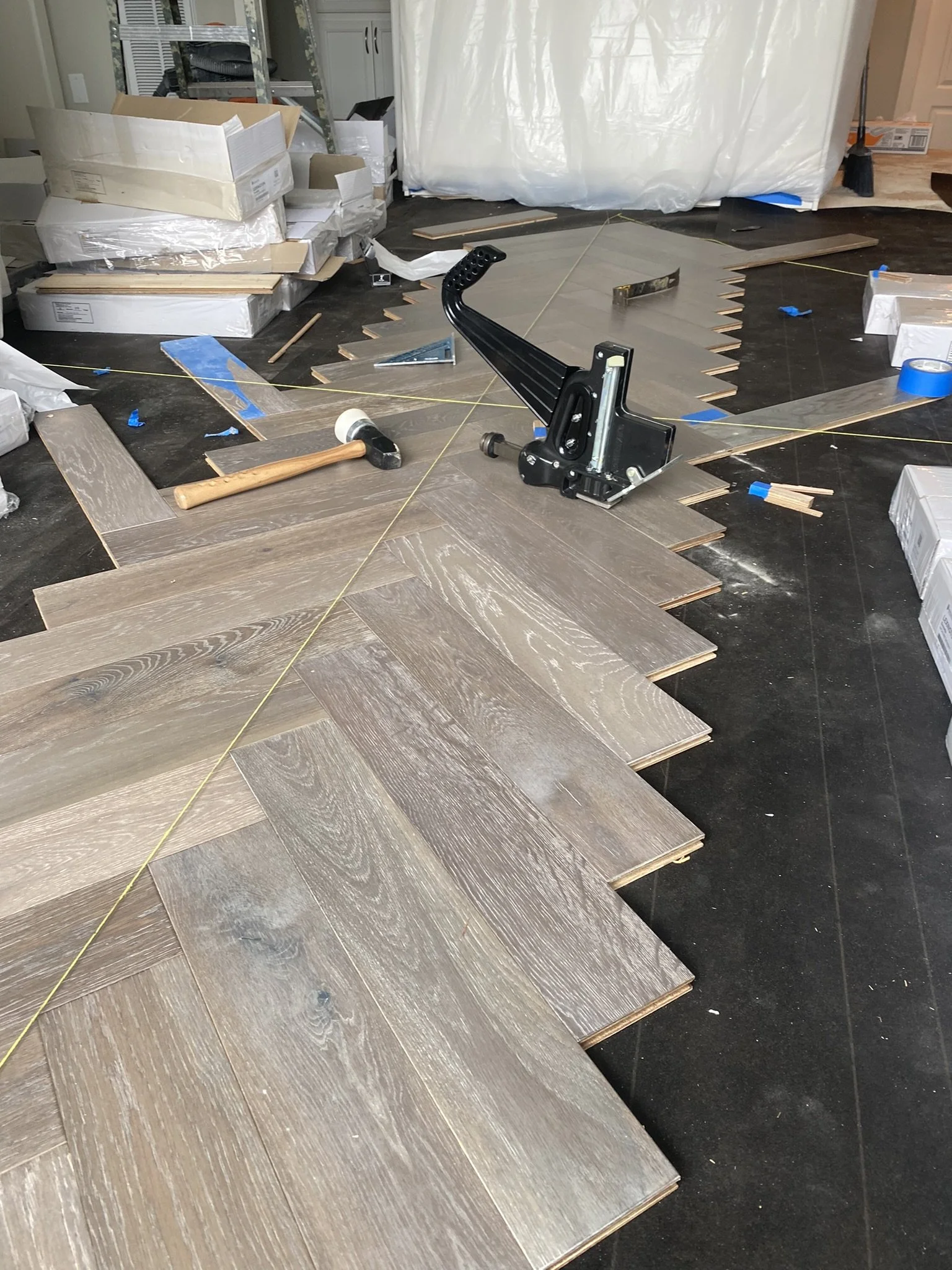 Herringbone wood floor installation in progress with tools and materials scattered around, including a mallet and flooring stapler.