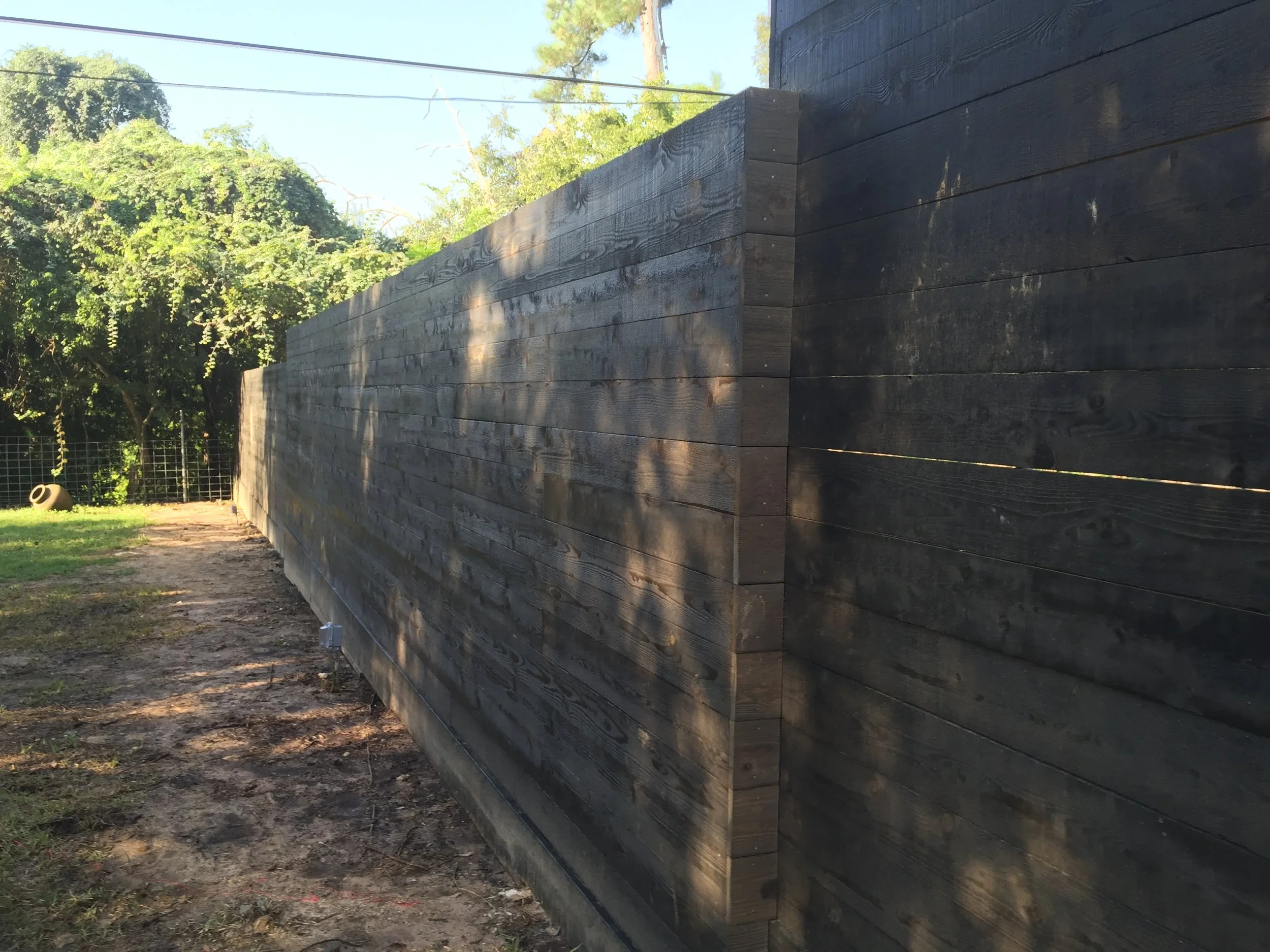 Long, dark wooden fence near yard with greenery in background