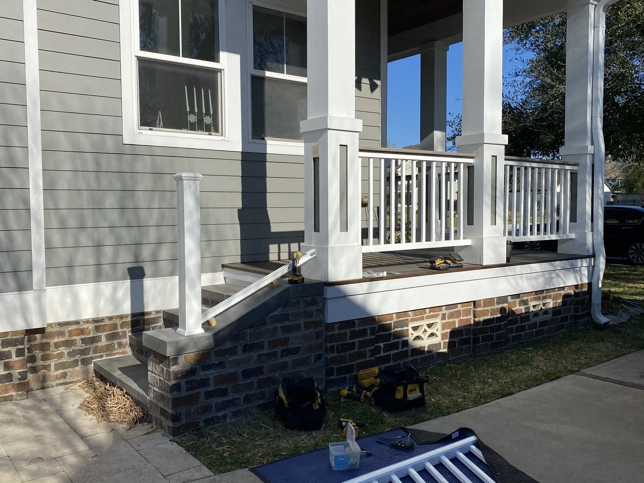 Front porch with white railing installation in progress, gray siding, brick foundation, construction tools on steps, and a grassy yard in sunlight.