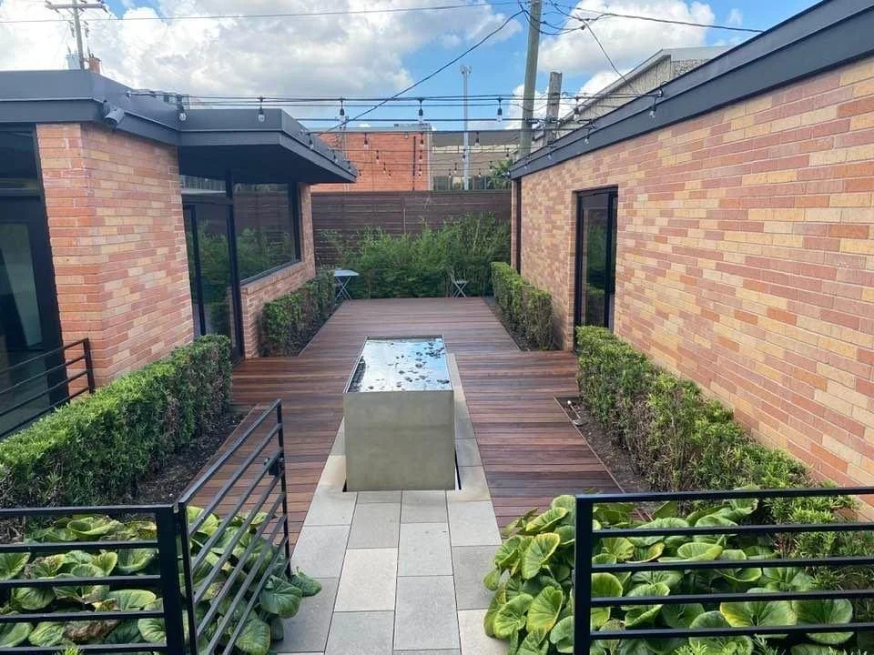 Modern outdoor courtyard with wooden decking, a rectangular water feature, and surrounded by brick walls and greenery.