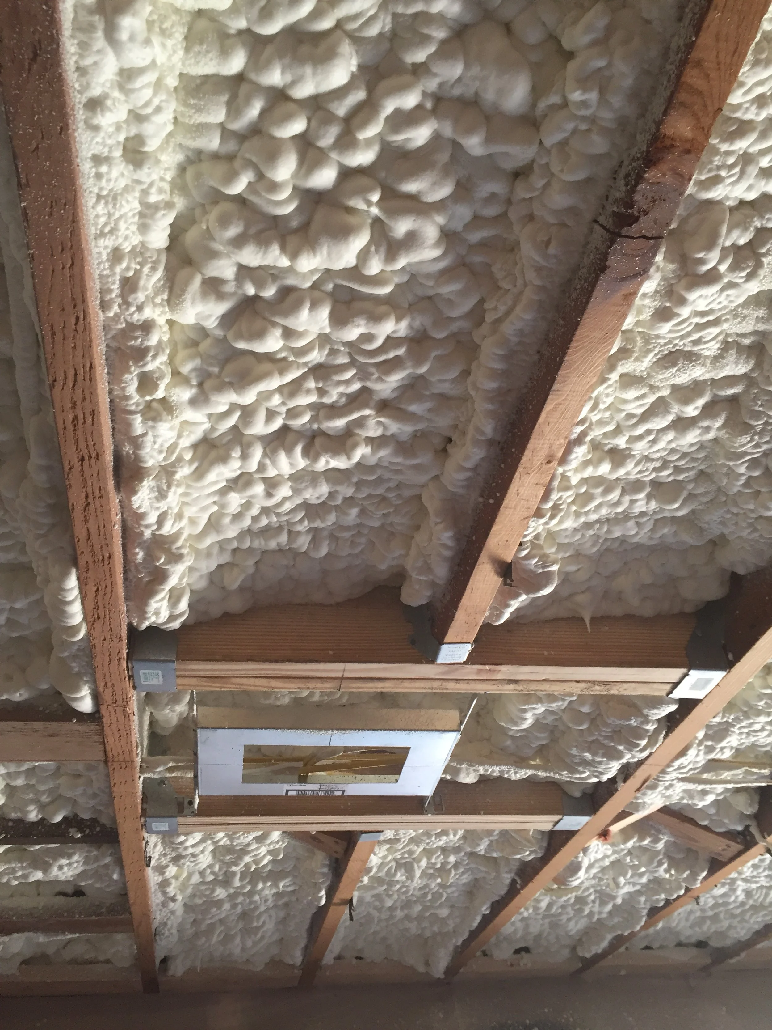 Spray foam insulation applied to ceiling with exposed wooden beams and a metal ventilation duct.