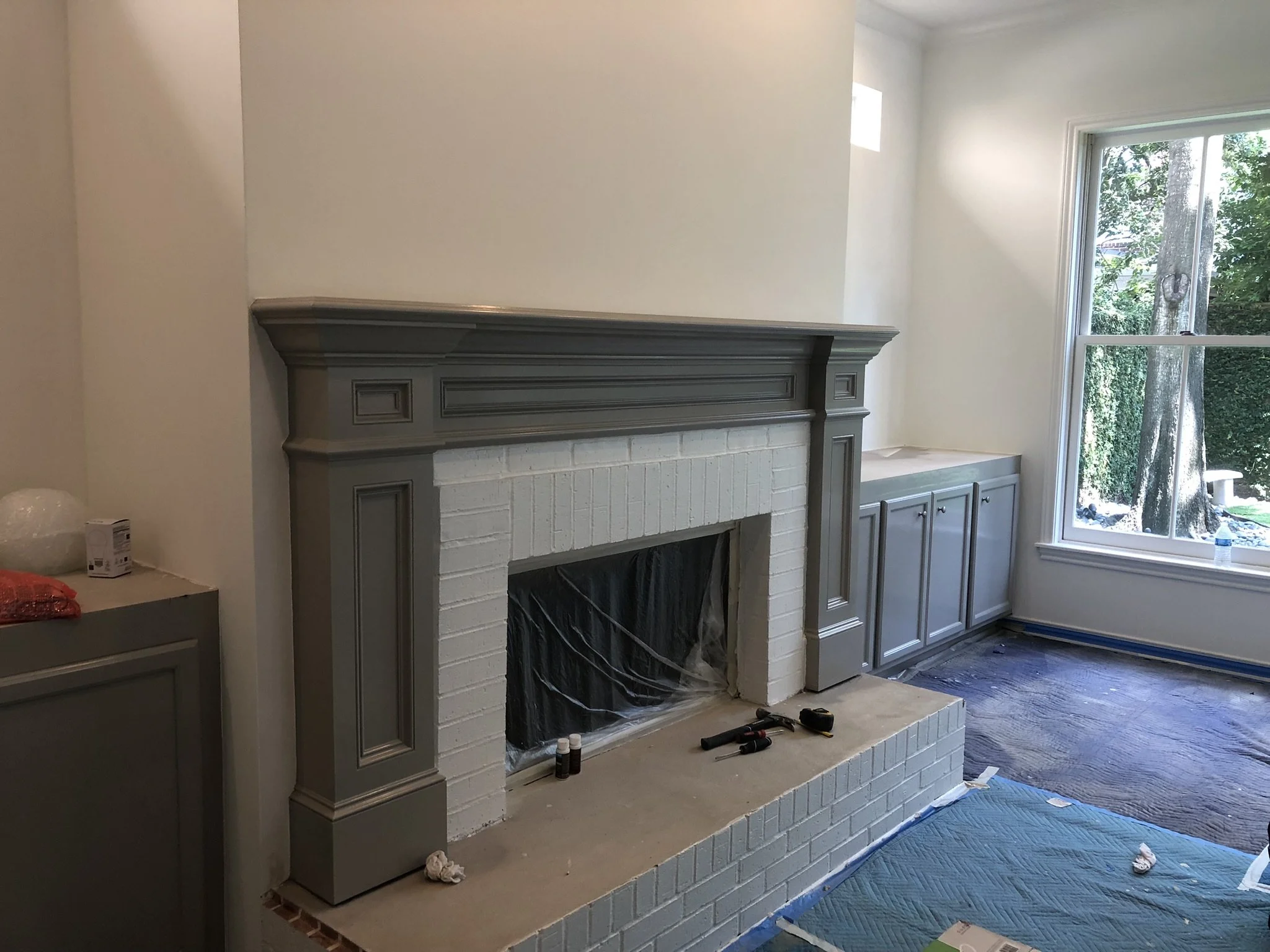 Living room with gray-painted fireplace and cabinetry, white walls, partially covered wood flooring, and large window.