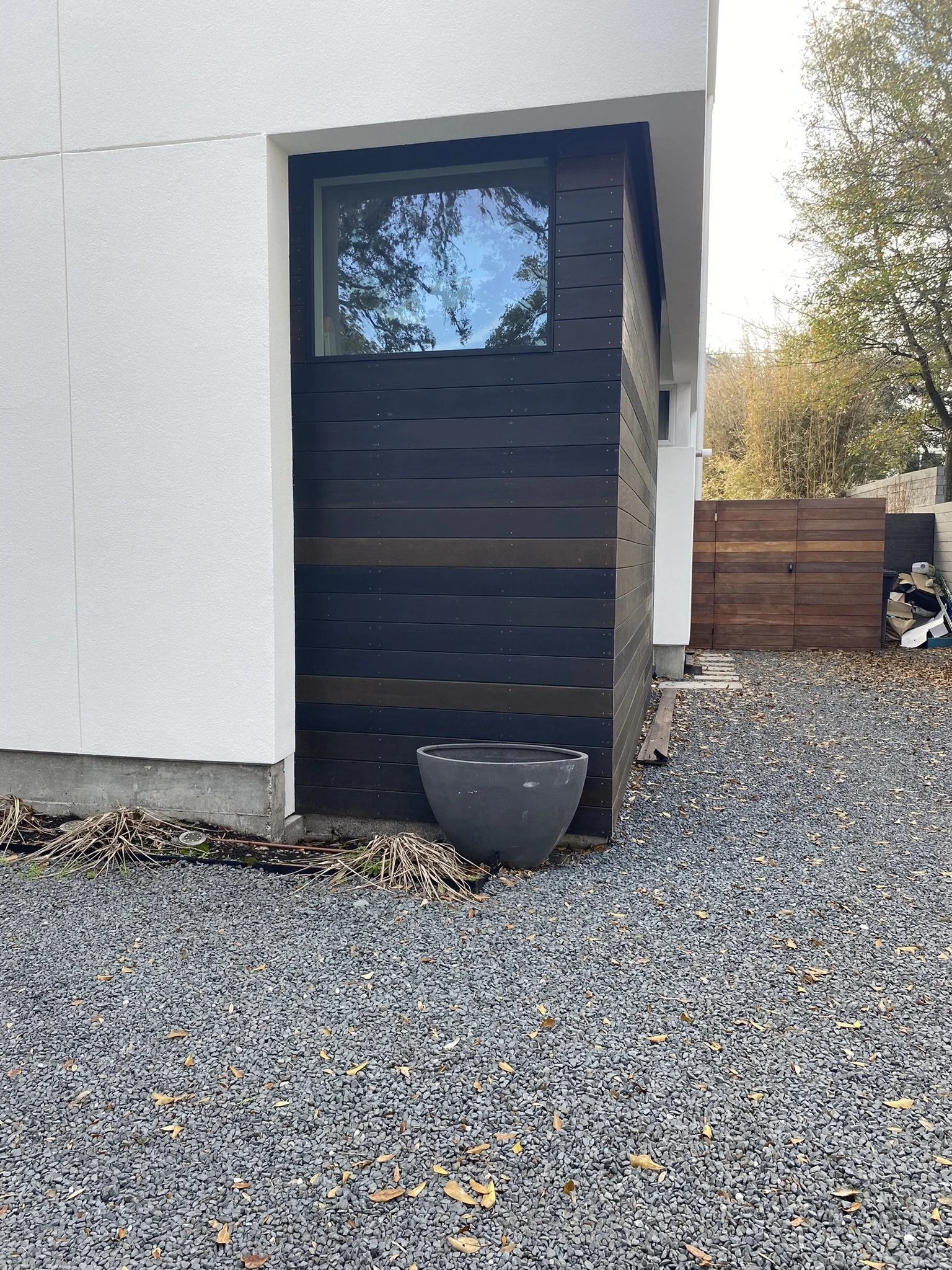 Modern house exterior with wood paneling, large window, empty planter, gravel ground, and scattered leaves.