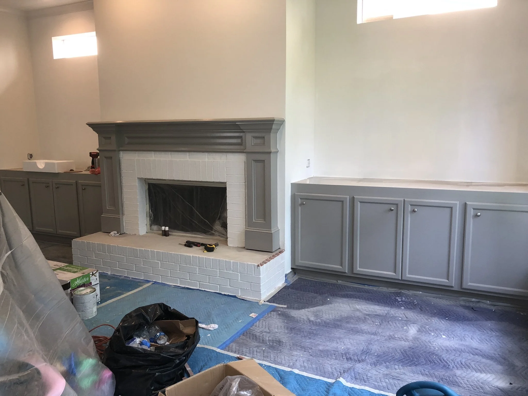 Room under renovation with a white brick fireplace, gray mantel and cabinetry, and covered floor. Tools and paint supplies are visible.