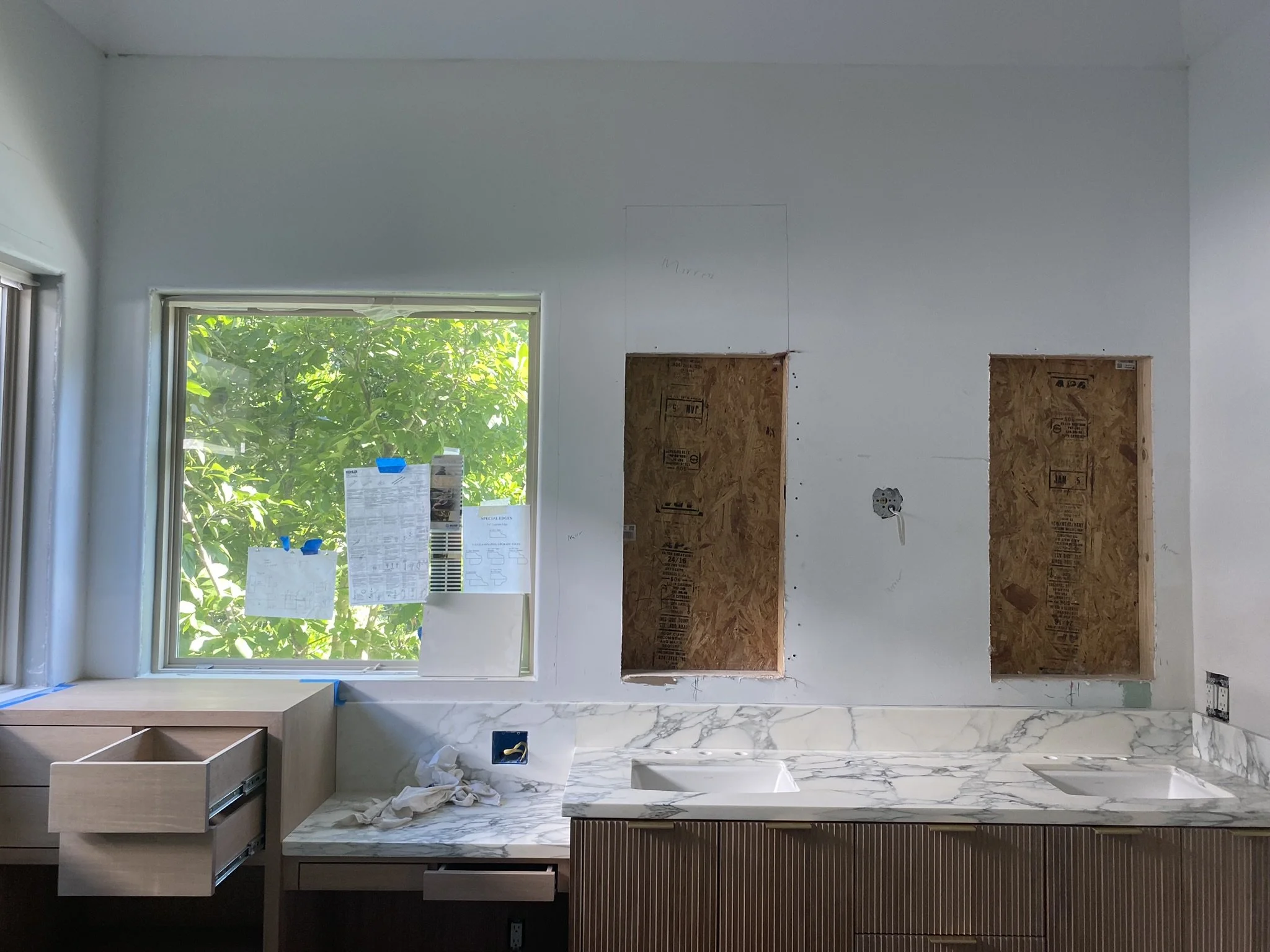 Partially renovated bathroom interior with marble countertop, open drawers, two rectangular wall cutouts for mirrors, and a large window with paperwork taped to it.