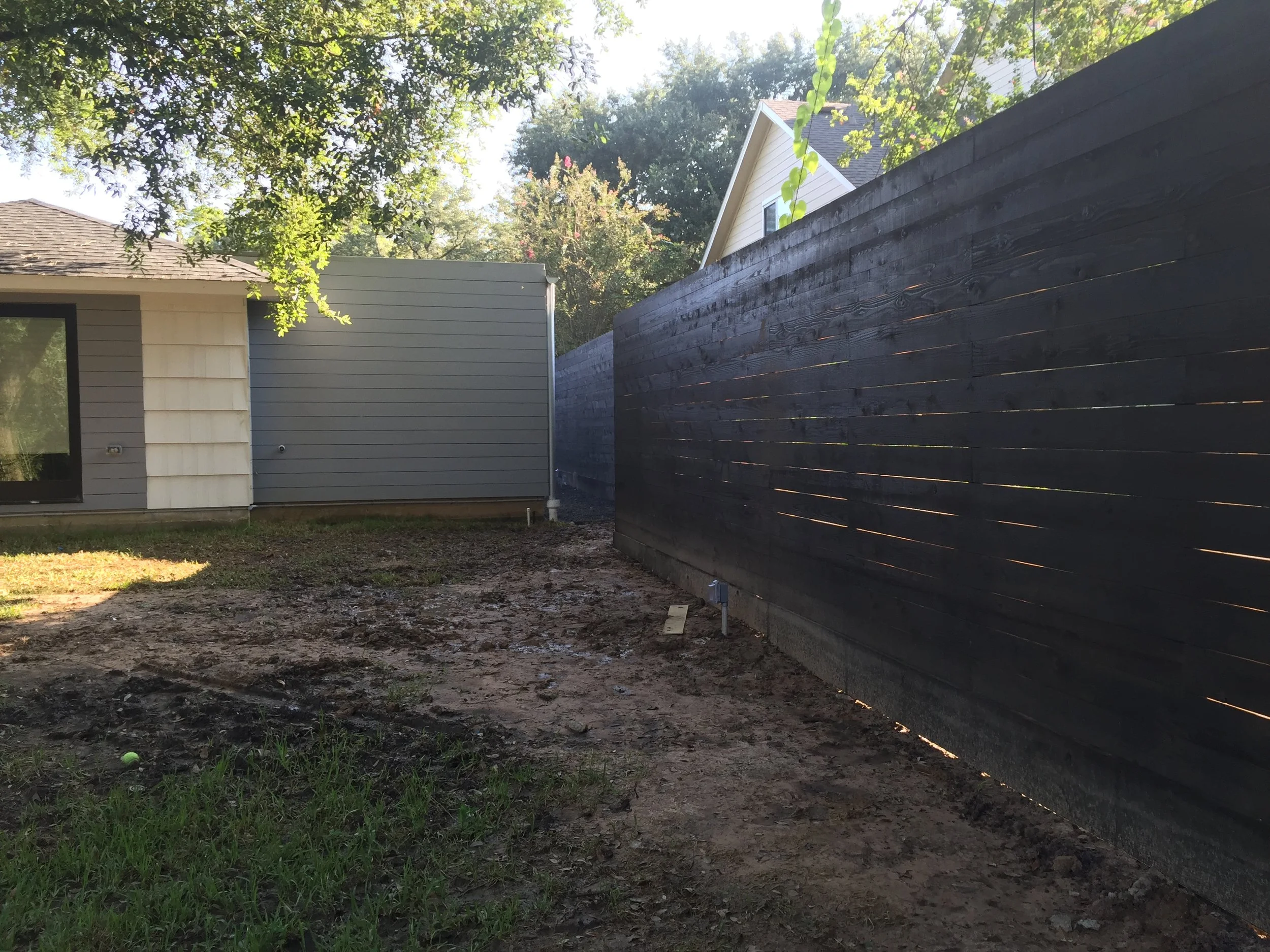 Backyard with a wooden fence and a house exterior, featuring siding and roofing, surrounded by trees.