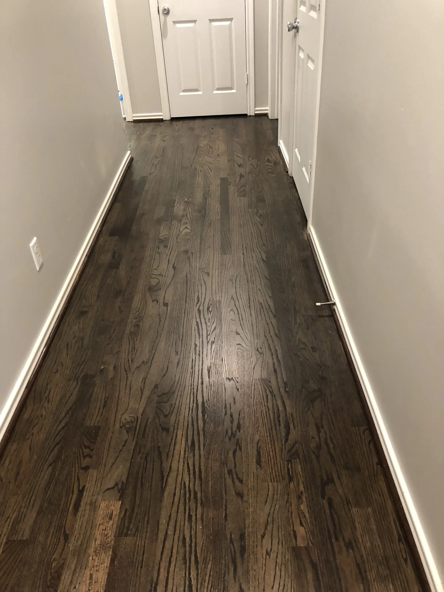 Narrow hallway with dark wood flooring, white walls, and white doors at the end and side.