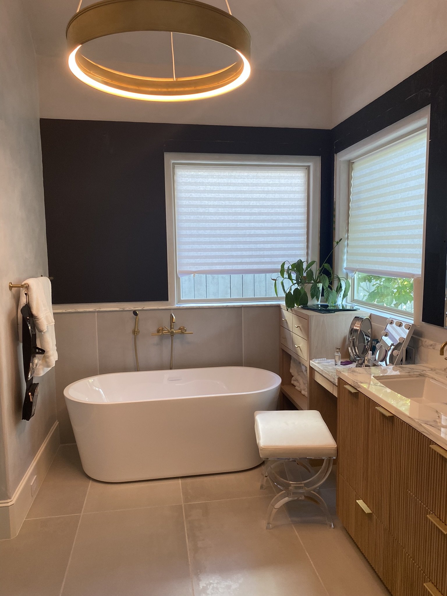 Modern bathroom with freestanding bathtub, stylish lighting, wooden vanity, upholstered stool, and indoor plants, featuring a neutral color palette.