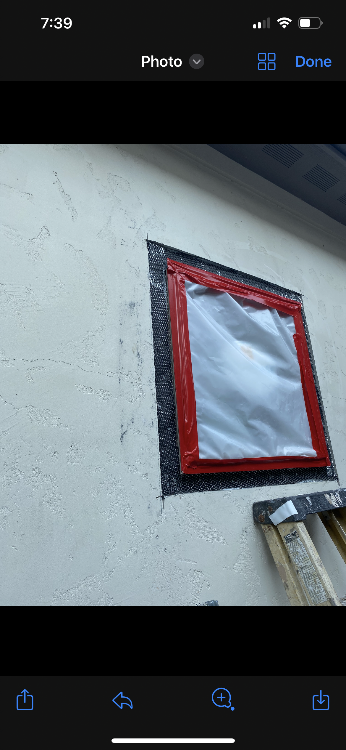Exterior wall with a window opening covered in plastic secured by red tape, surrounded by a metal mesh frame, and a ladder leaning against the wall.