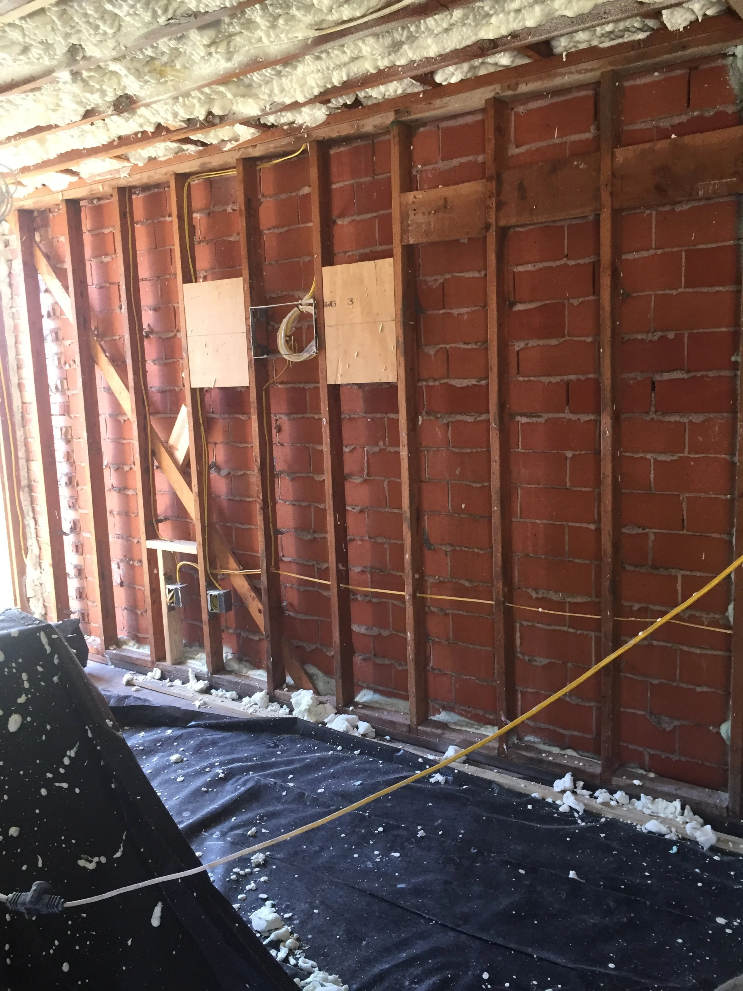 Exposed brick wall with wooden studs and foam insulation, showing wiring and construction materials.