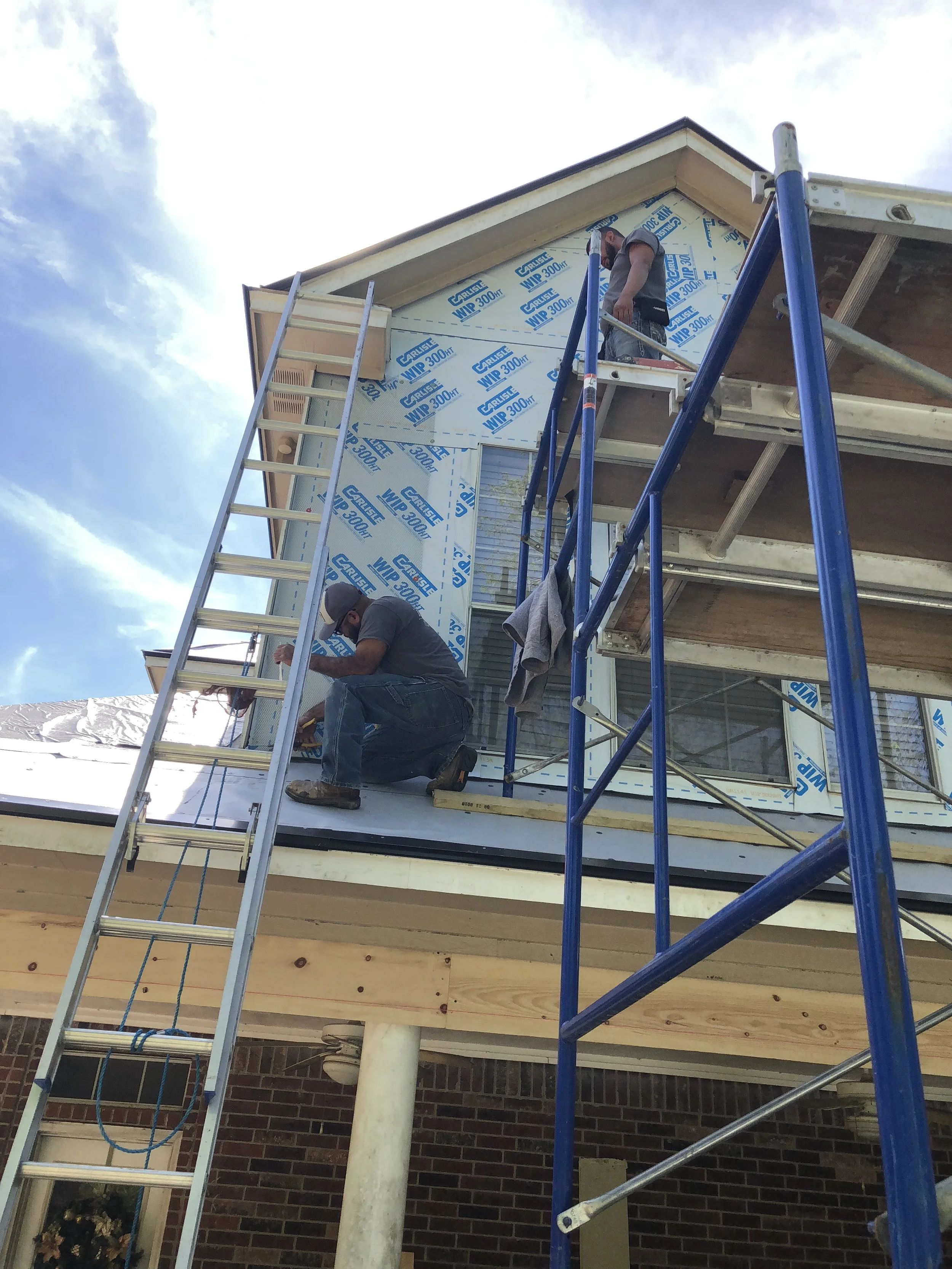 Construction workers on scaffolding and ladders installing house wrap on a building's roof and wall.