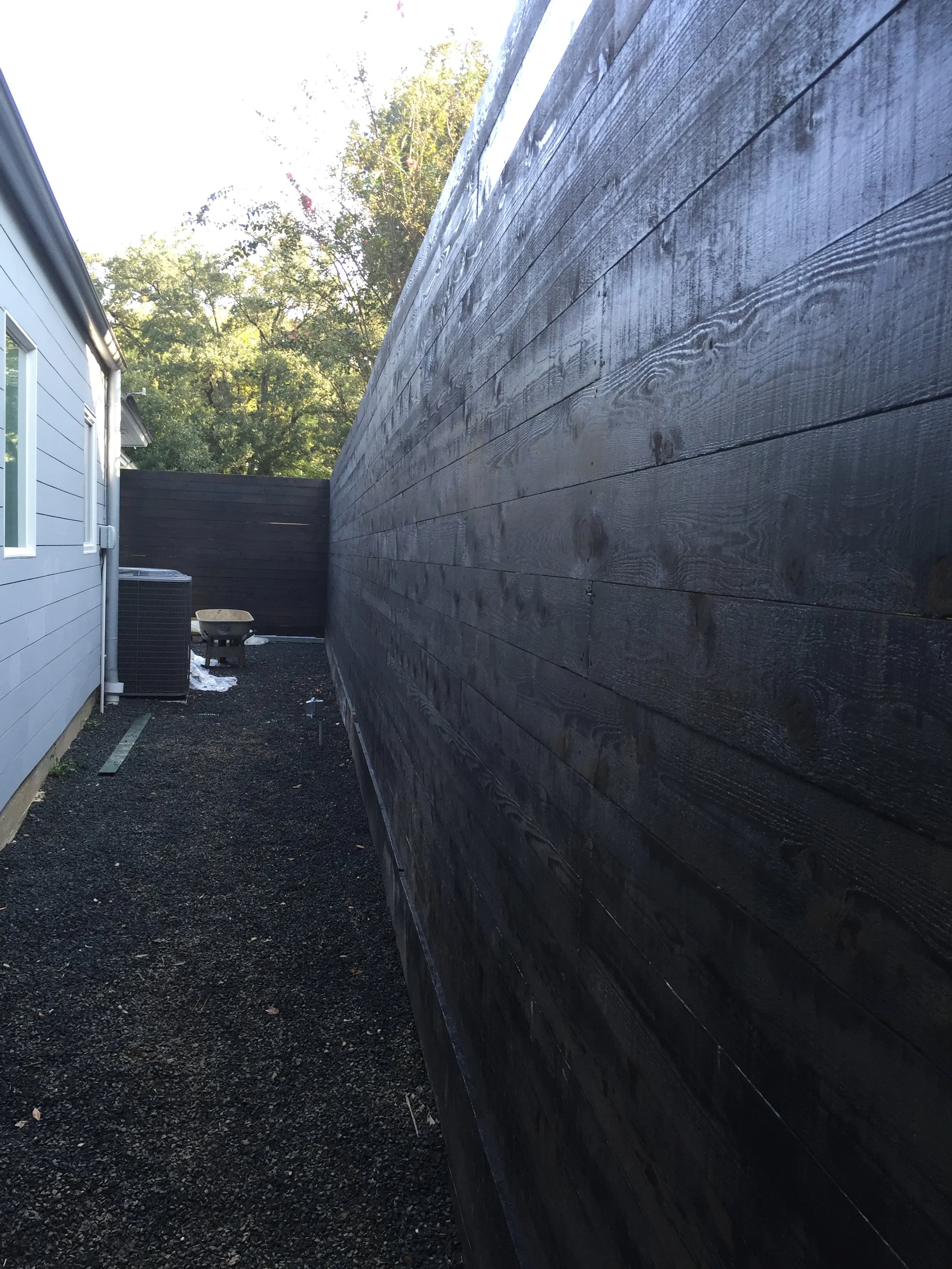 Narrow alleyway between a gray building and a tall wooden fence, with gravel on the ground and an outdoor air conditioning unit visible.