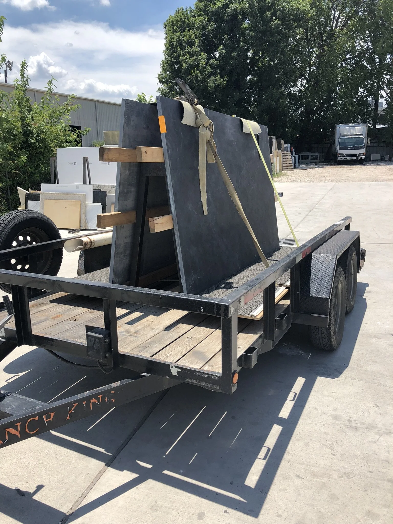 A flatbed trailer carrying large vertically-oriented slabs of stone or concrete, secured with straps, parked on a driveway near a commercial building and trees.