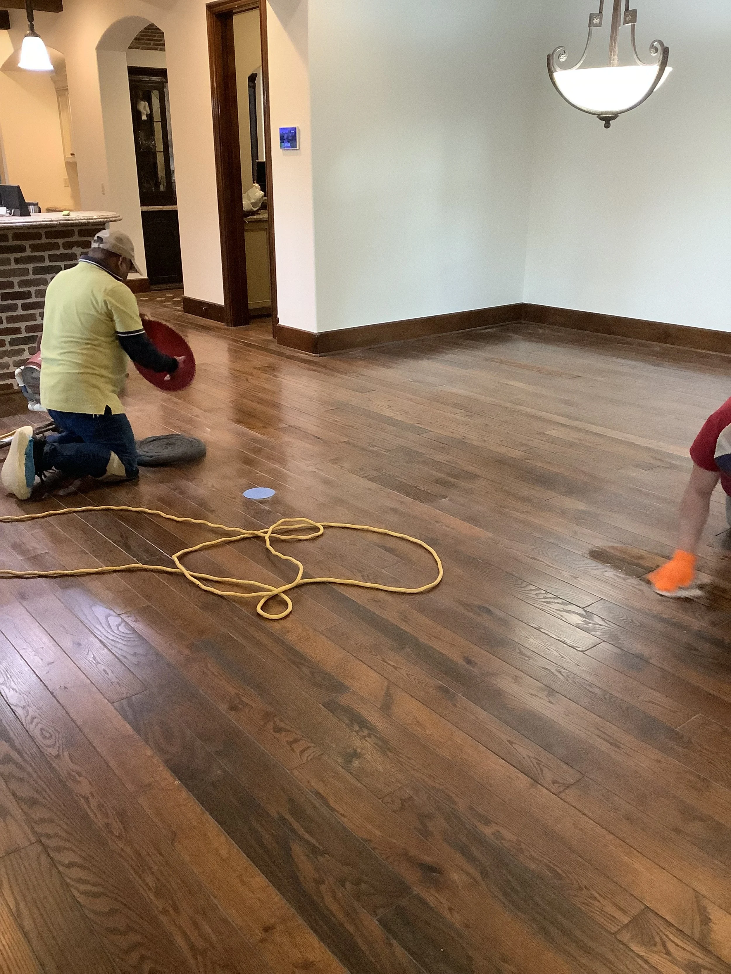 Two people cleaning and polishing a hardwood floor in a residential interior, with visible electrical cord and modern chandelier.