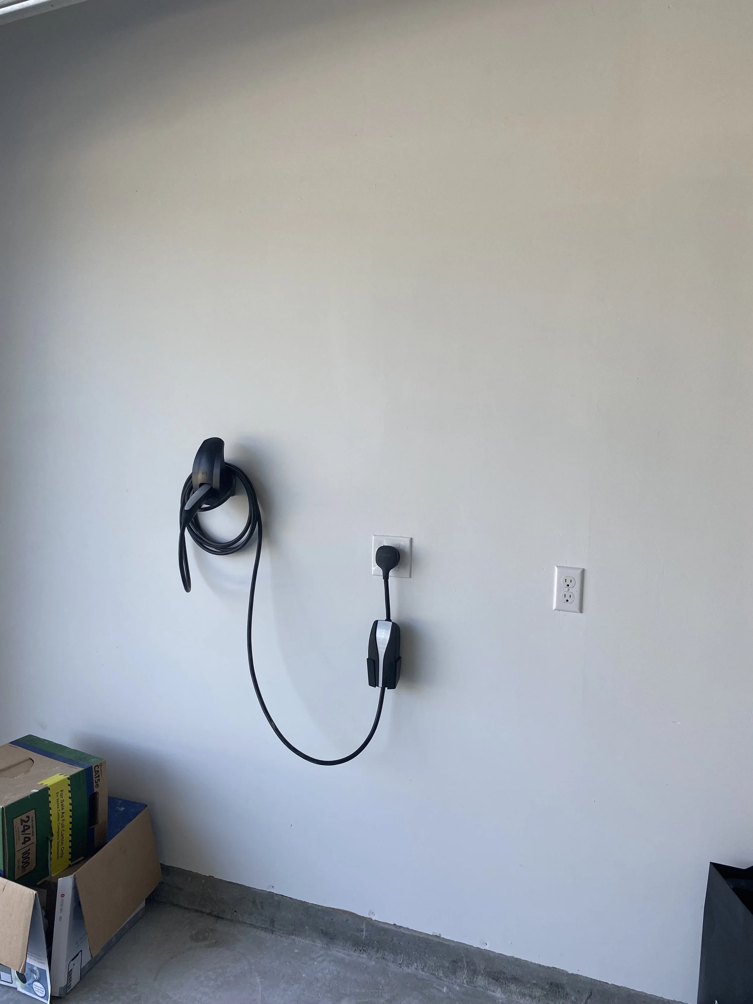 Garage wall with an electric vehicle (EV) charging station and cable. Cardboard boxes are stacked on the floor nearby.