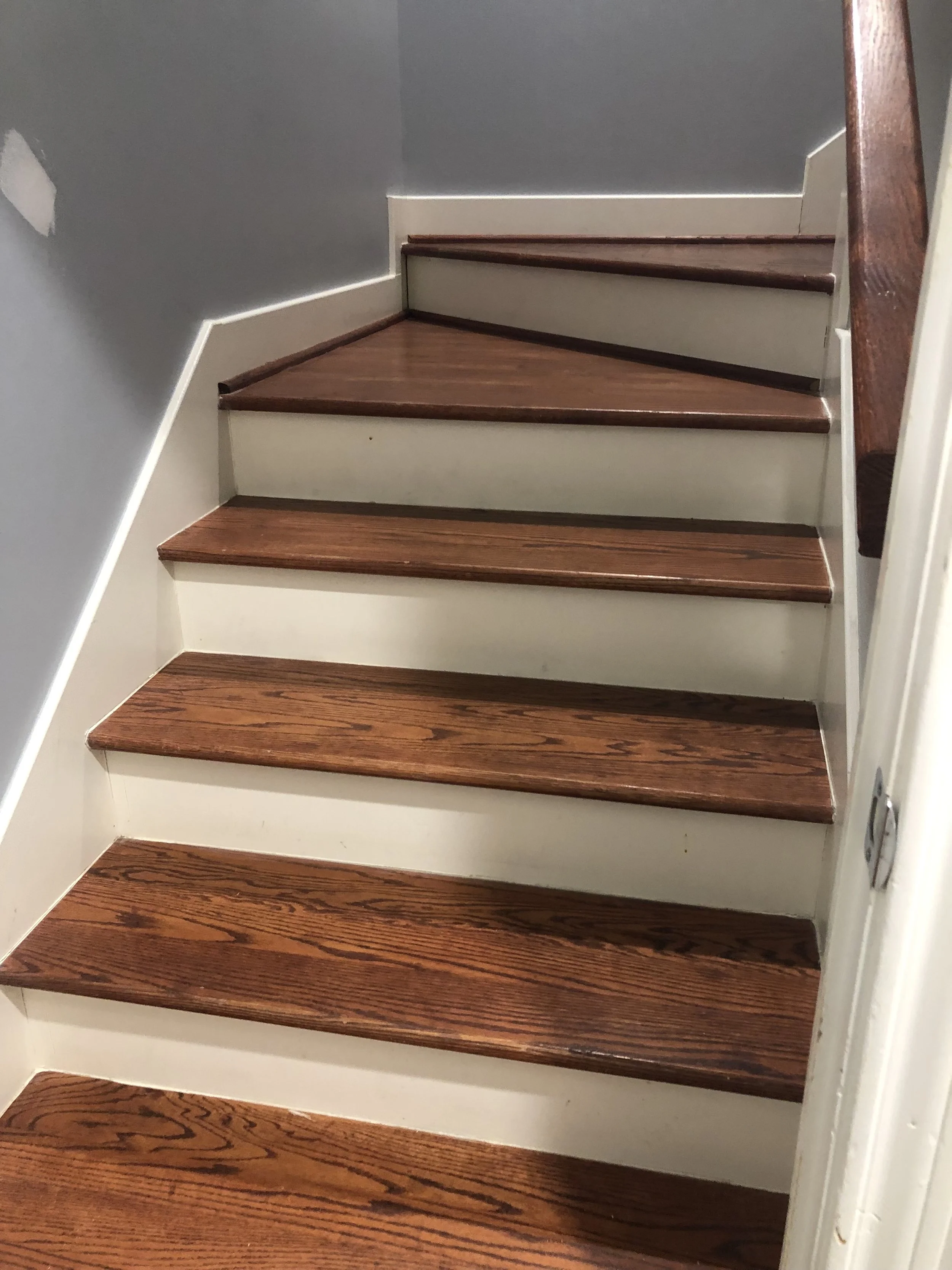 Wooden staircase with dark brown treads and white risers, leading upward with a turn to the right, against a gray wall.