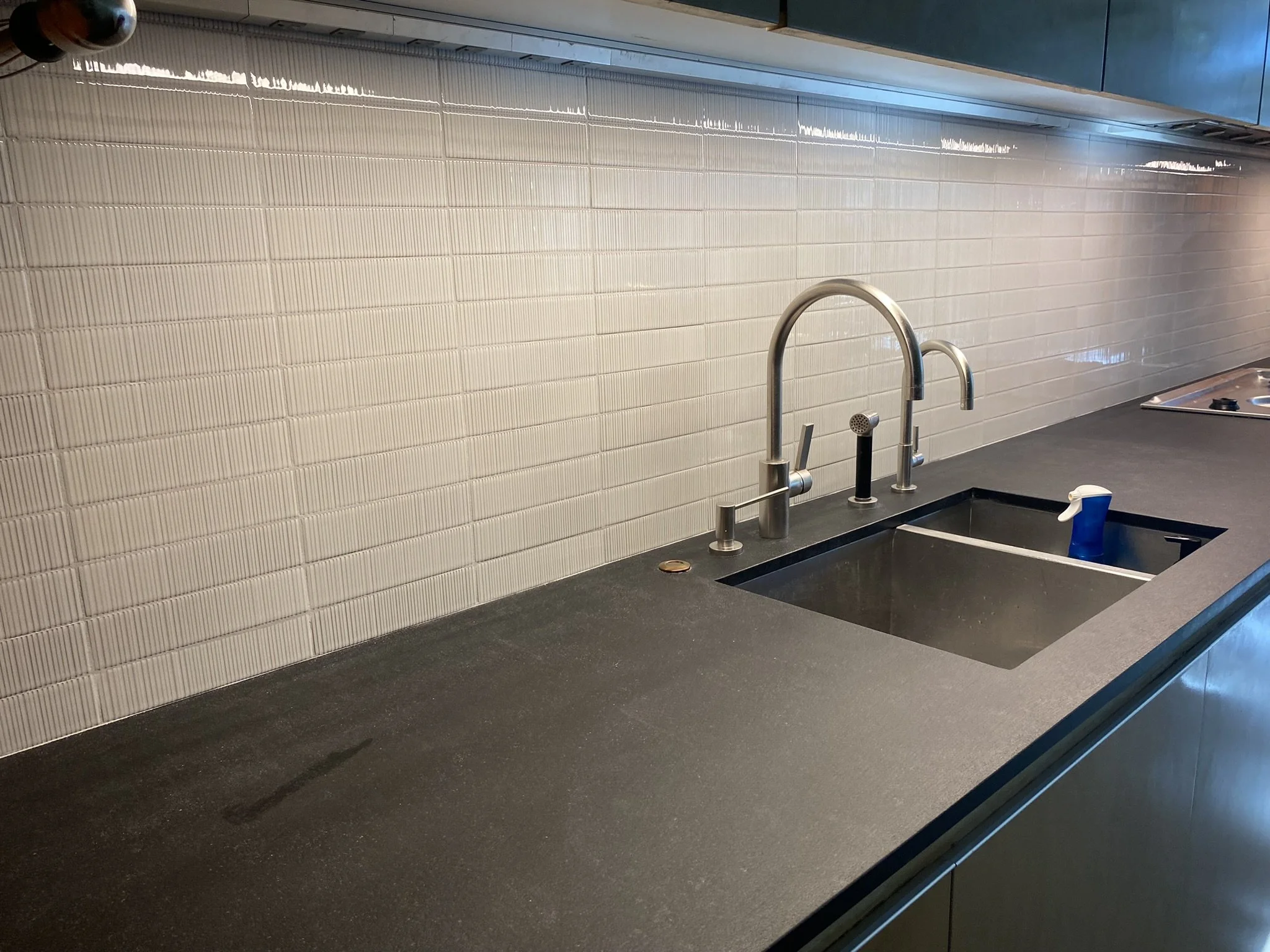 Modern kitchen countertop with a dark surface, featuring a stainless steel sink and faucet, a textured white tile backsplash, and a spray bottle.