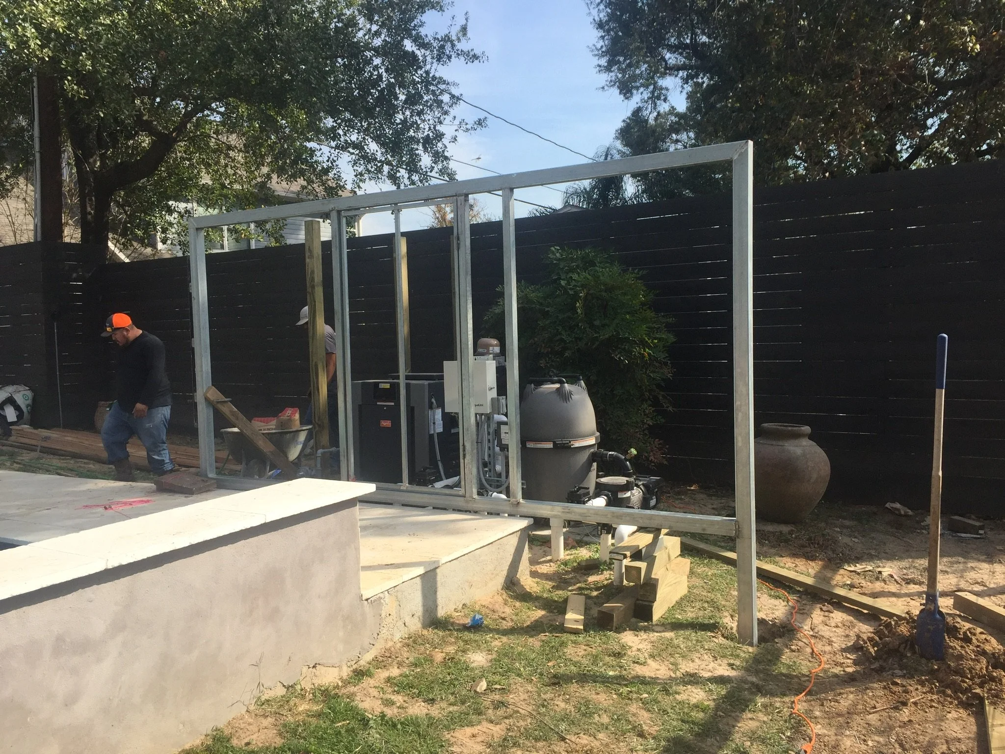 Outdoor construction site with metal framework, pool equipment, and a worker in an orange cap. A large pot is in the background next to a wooden fence and trees.