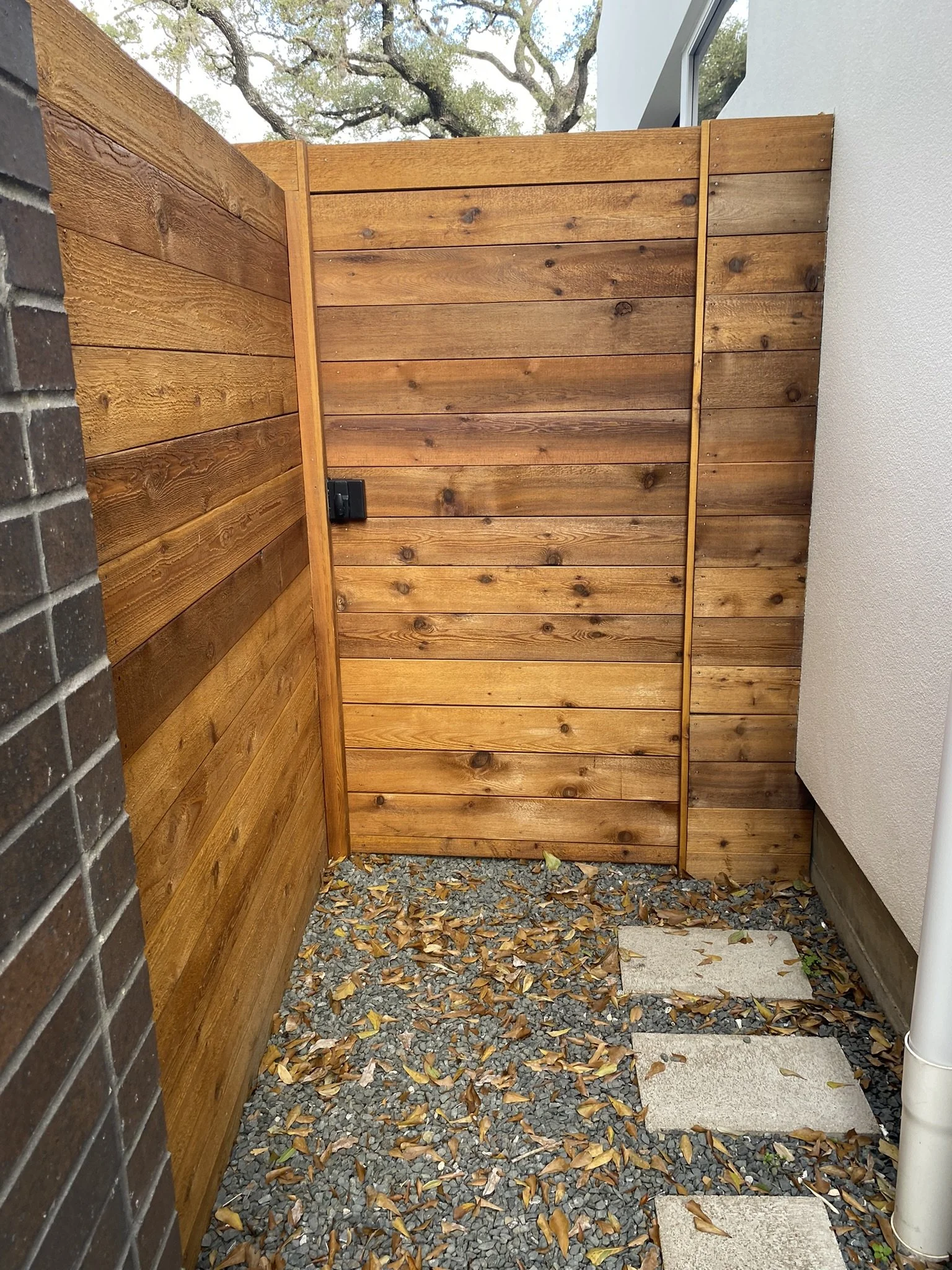 Wooden gate and fence with gravel and fallen leaves on the ground, next to a white building wall.