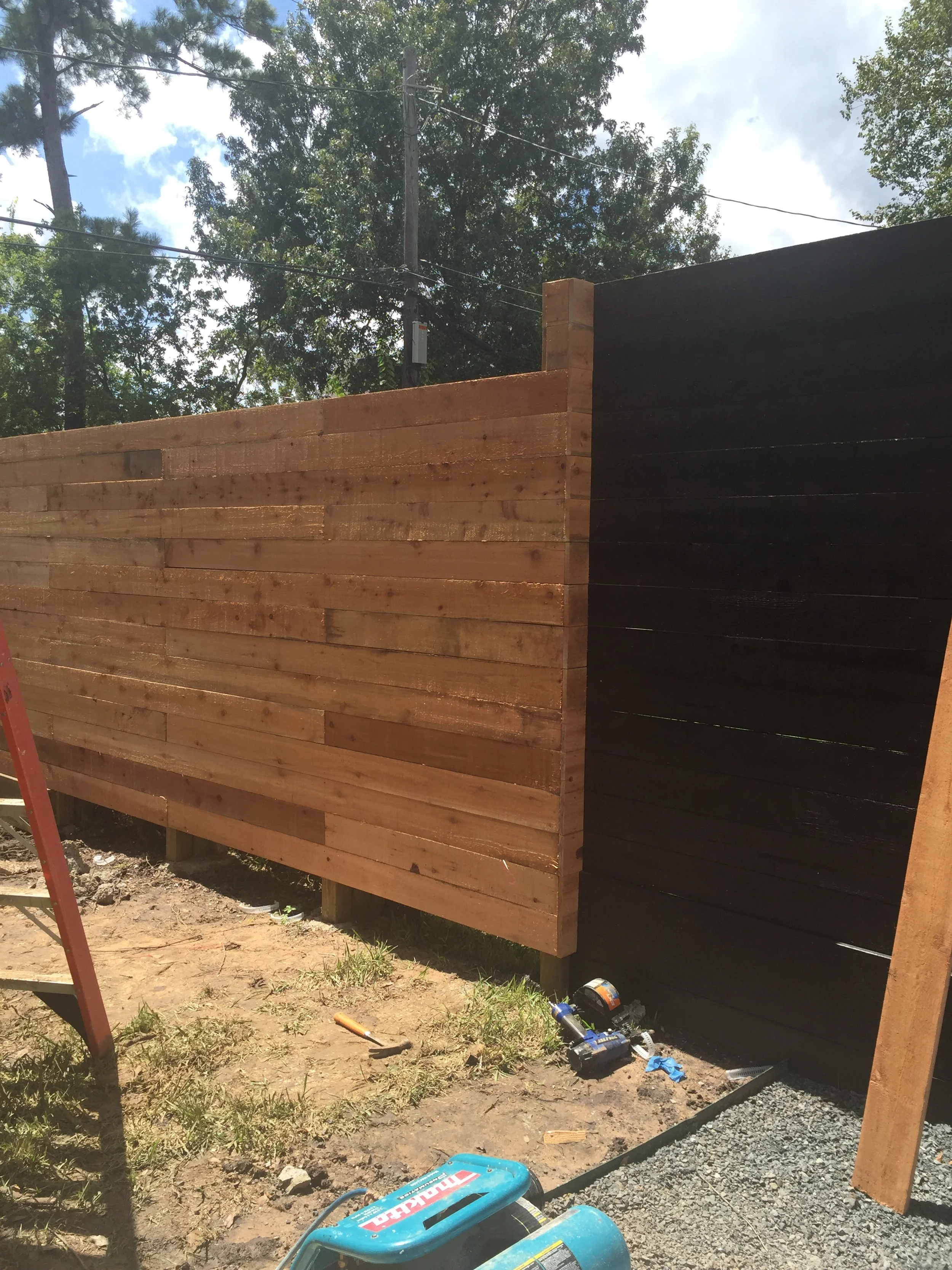 Wooden fence under construction with tools and a ladder nearby in a backyard setting, surrounded by trees under a partly cloudy sky.