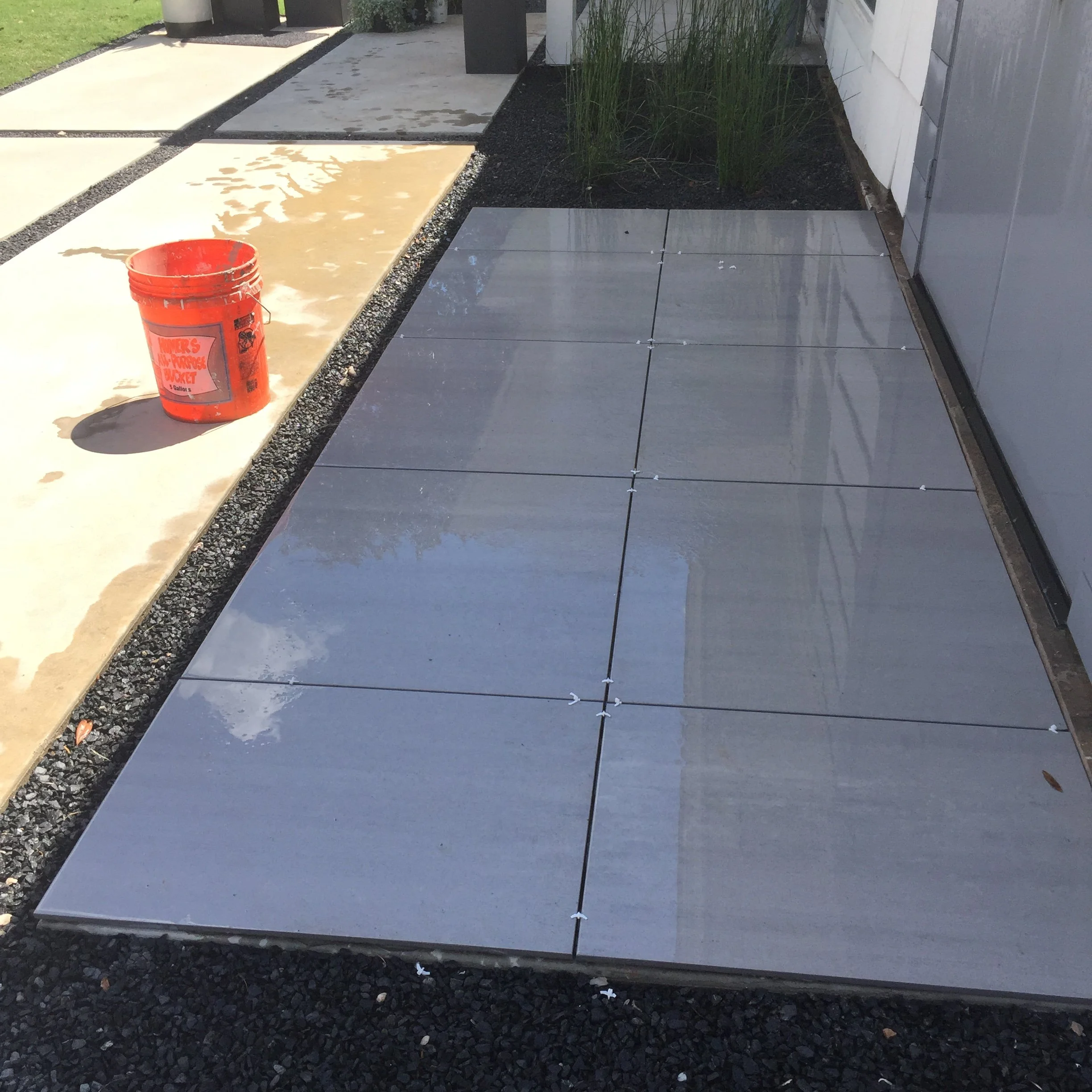 Gray tiles being installed on a patio with spacers, adjacent to a path and a red bucket.