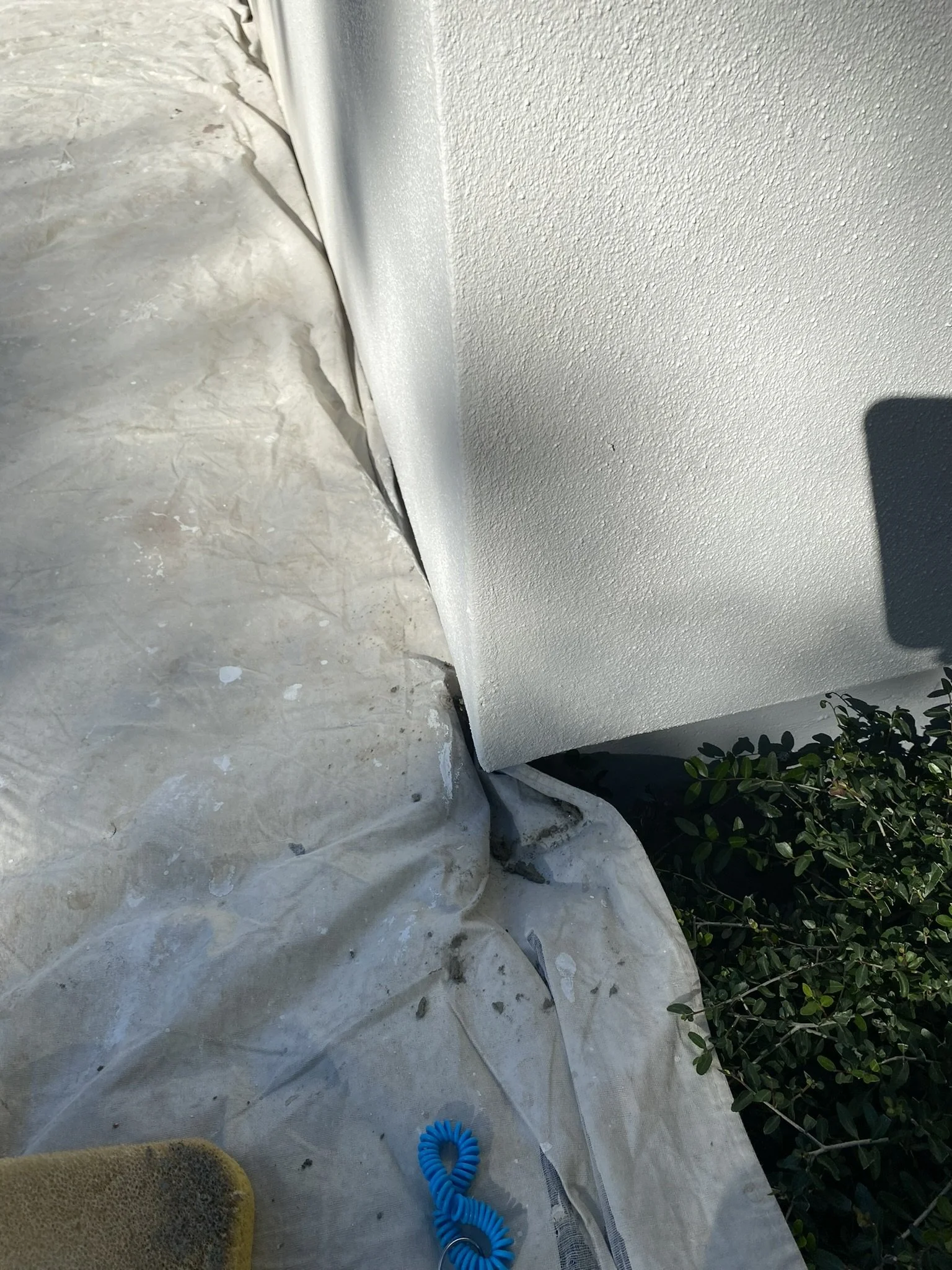 Corner of a building with white stucco walls, a tarp on the ground, and some greenery. A blue coiled cord and a sponge are on the tarp.