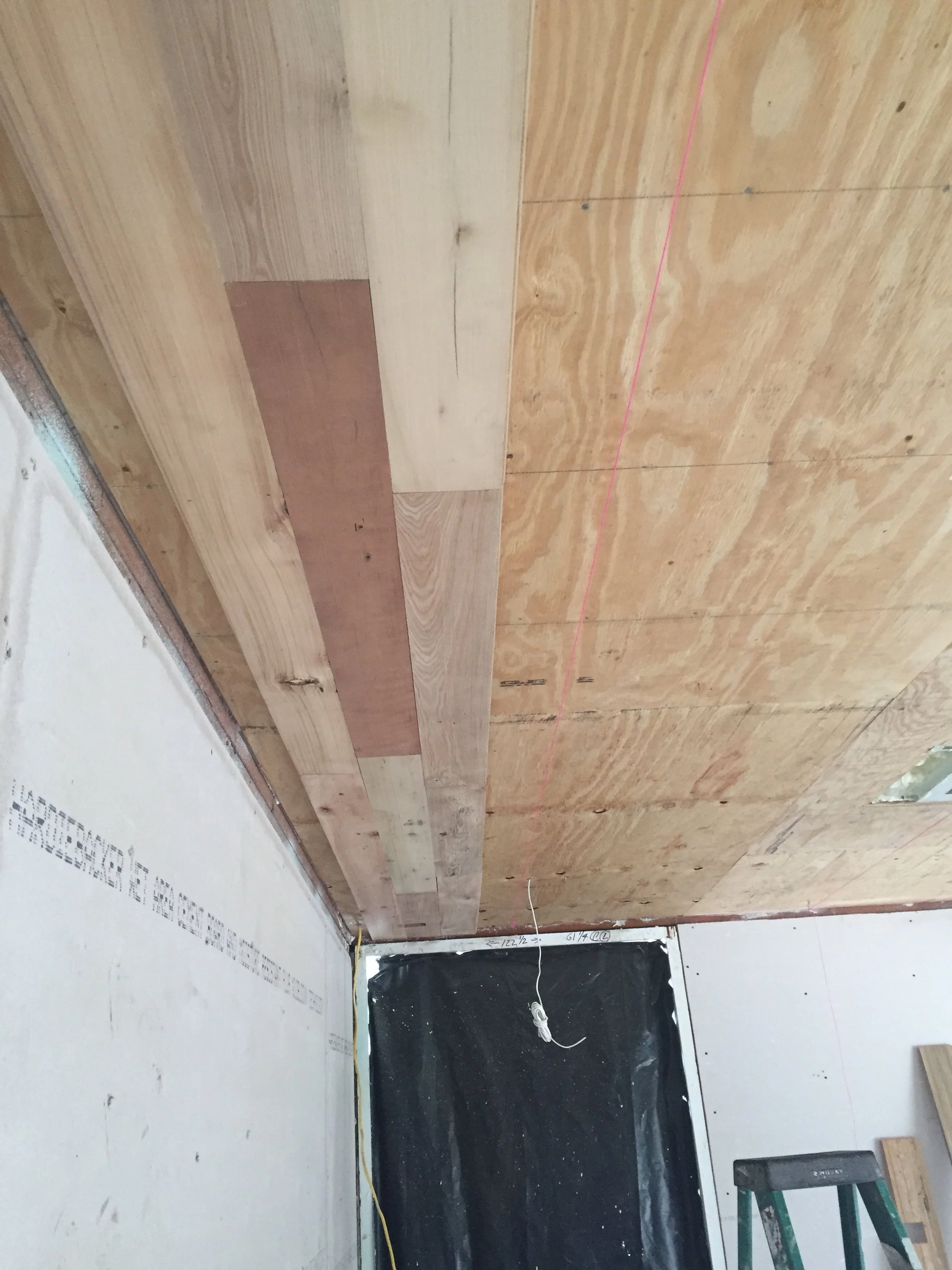 Room under construction with unfinished wooden ceiling panels, partially installed drywall, and a ladder.