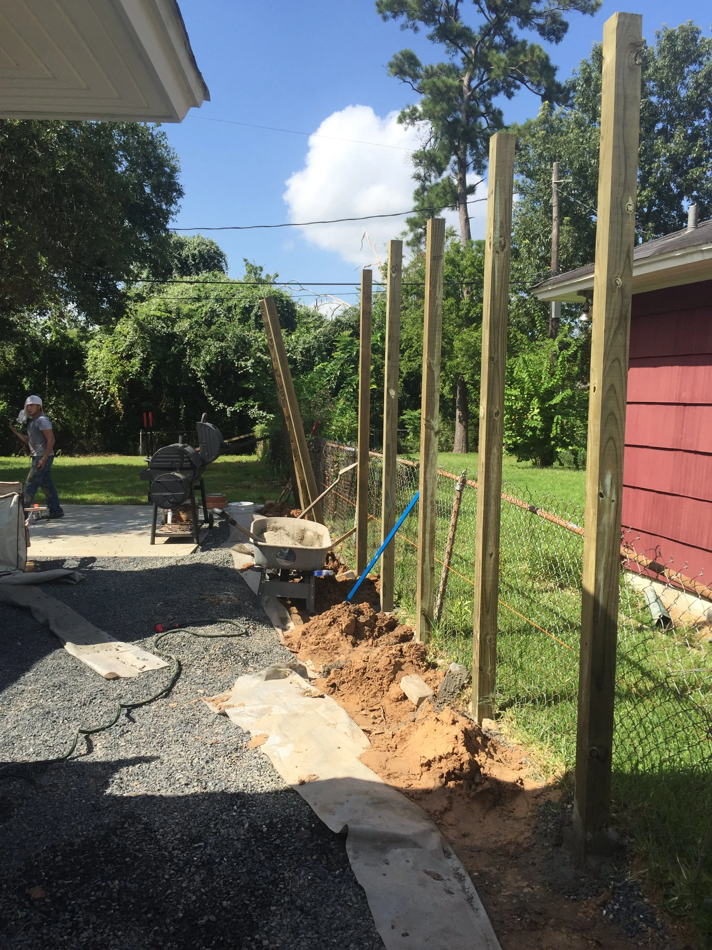 Backyard construction scene with wooden fence posts, wheelbarrow, and gravel path.