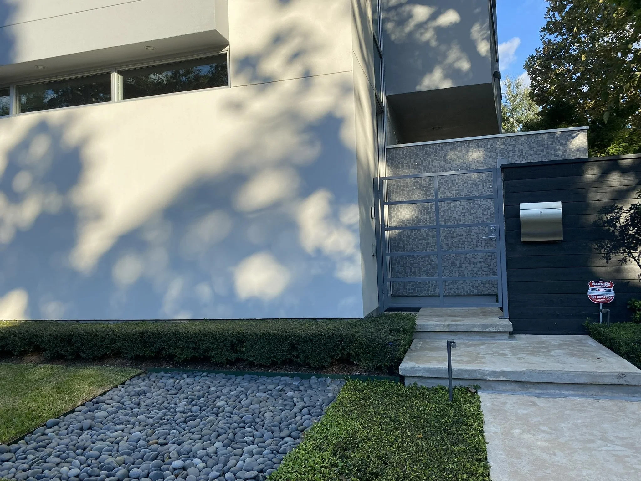 Modern house entrance with a minimalist design, featuring a white wall, a metal gate, landscaped greenery, a pathway with pebbles, and a security sign.