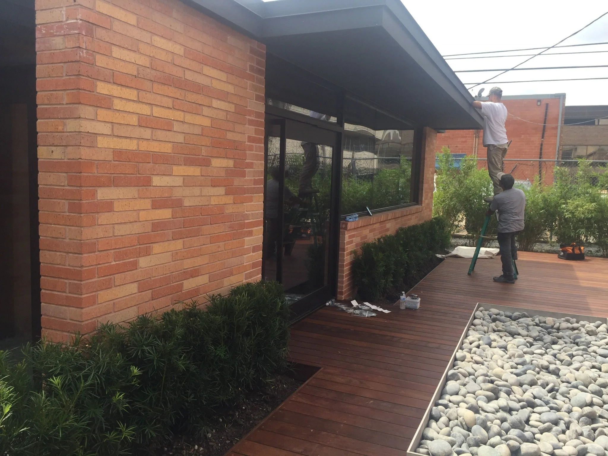 Two workers installing roofing on a brick building with a wooden deck and decorative stone area.