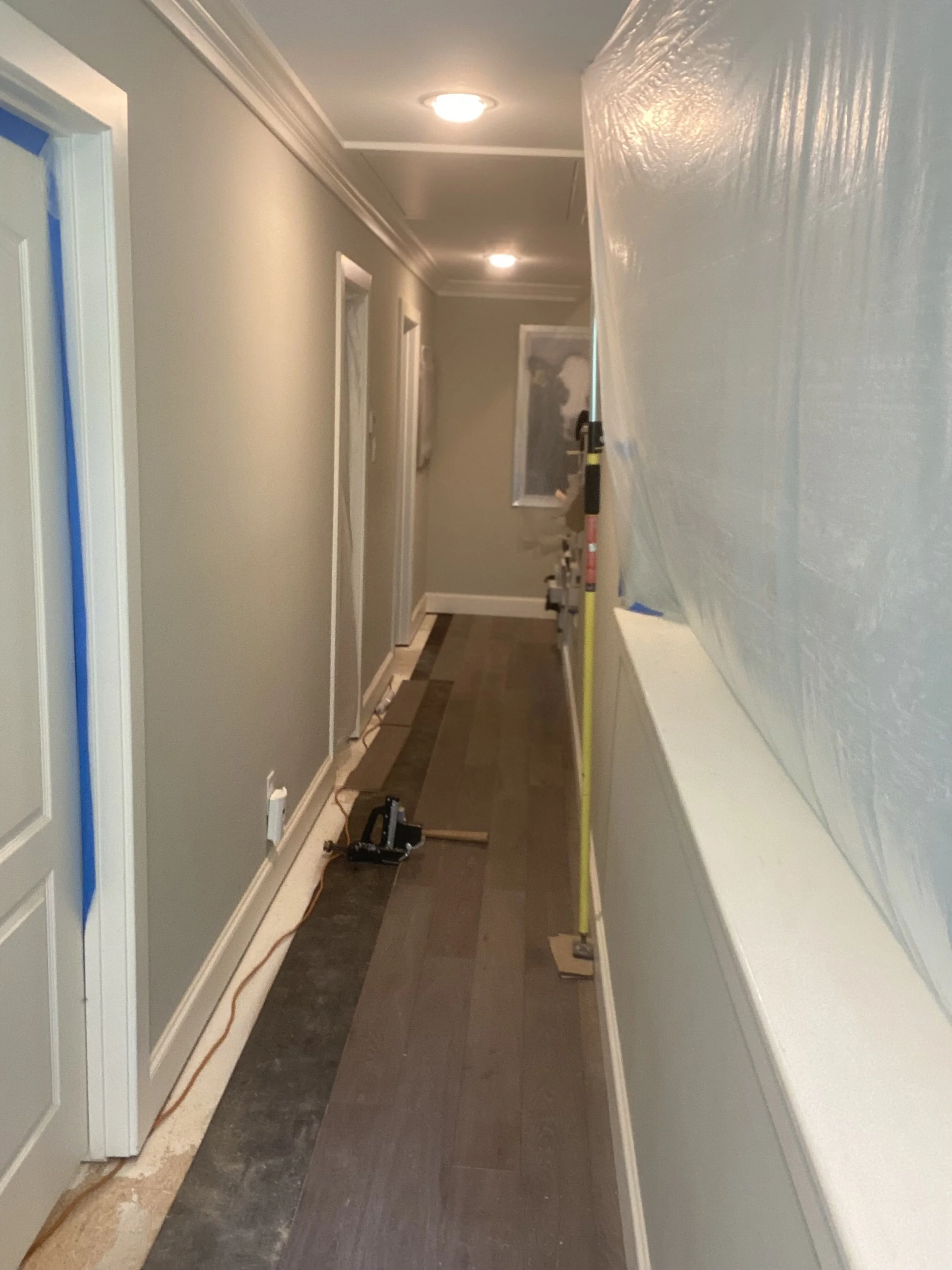 A hallway under renovation with gray walls, white doors, new flooring partially installed, plastic sheeting covering one side, and construction tools on the floor.